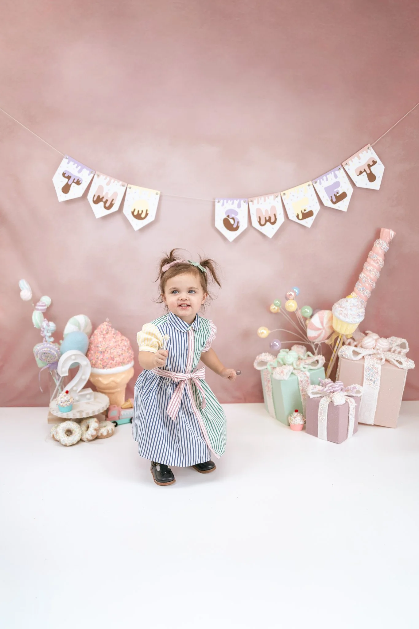 A young girl standing in front of colorful birthday decorations, including presents, balloons, and a cake, with a pink backdrop and a banner that reads 'Two'.