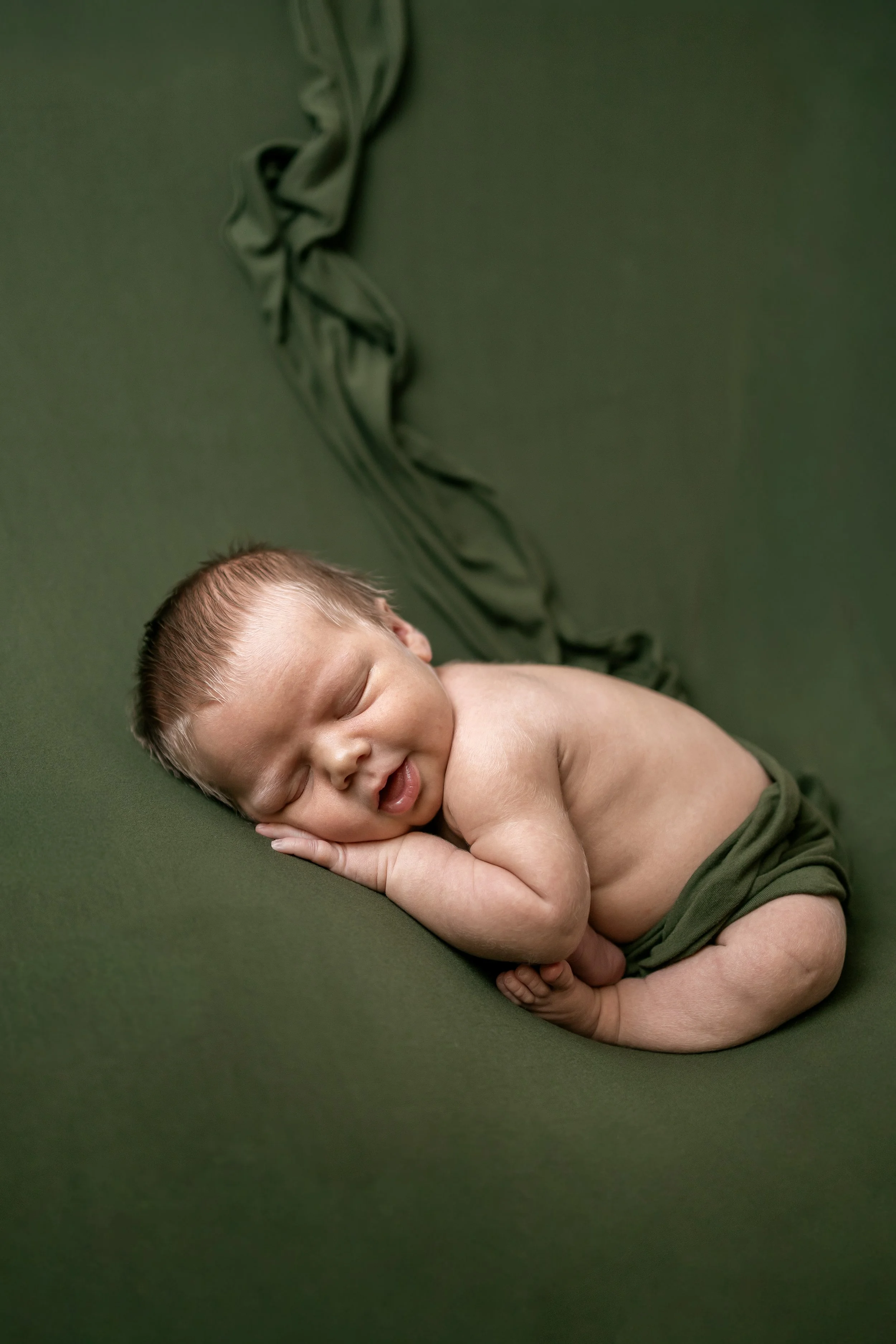A newborn baby peacefully sleeping on a green blanket with a matching green drape in the background.