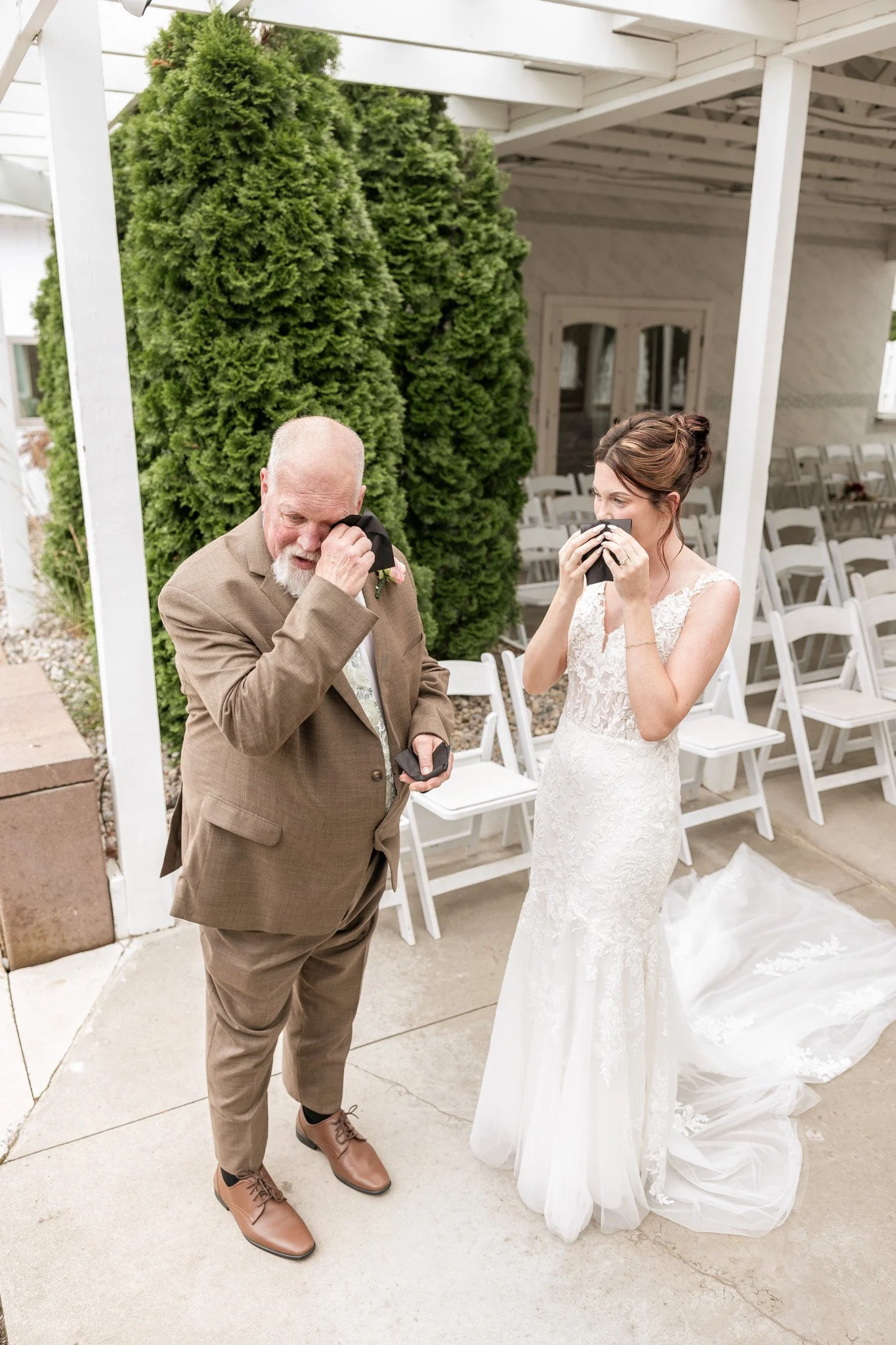 A bride in a white wedding dress and a man in a brown suit are emotional, wiping tears in an outdoor wedding setting. Rows of chairs are behind them, and a white pergola and greenery are also visible.