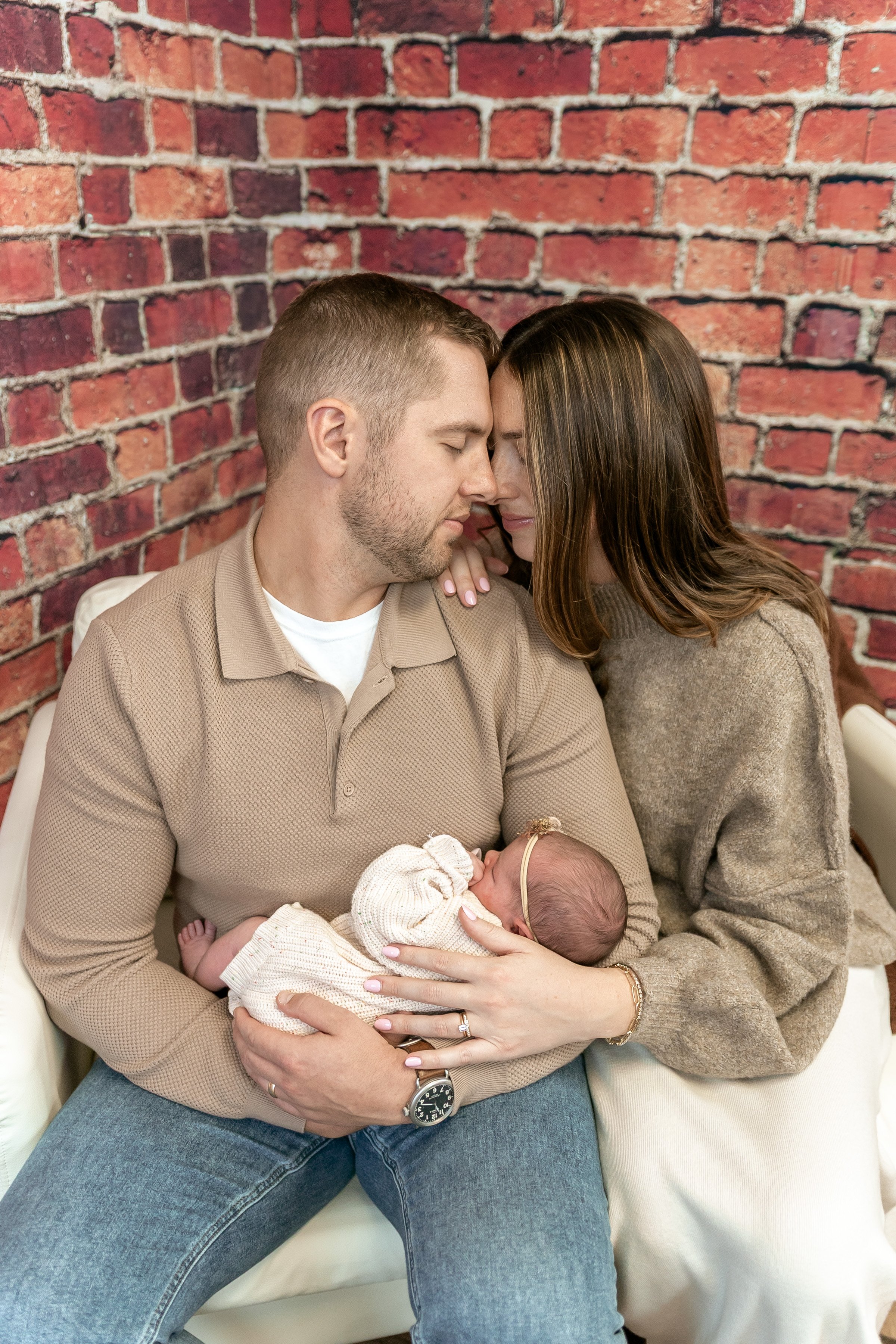 A couple holding their newborn baby in front of a brick wall, with the couple touching foreheads and the woman resting her hand on the man's shoulder.