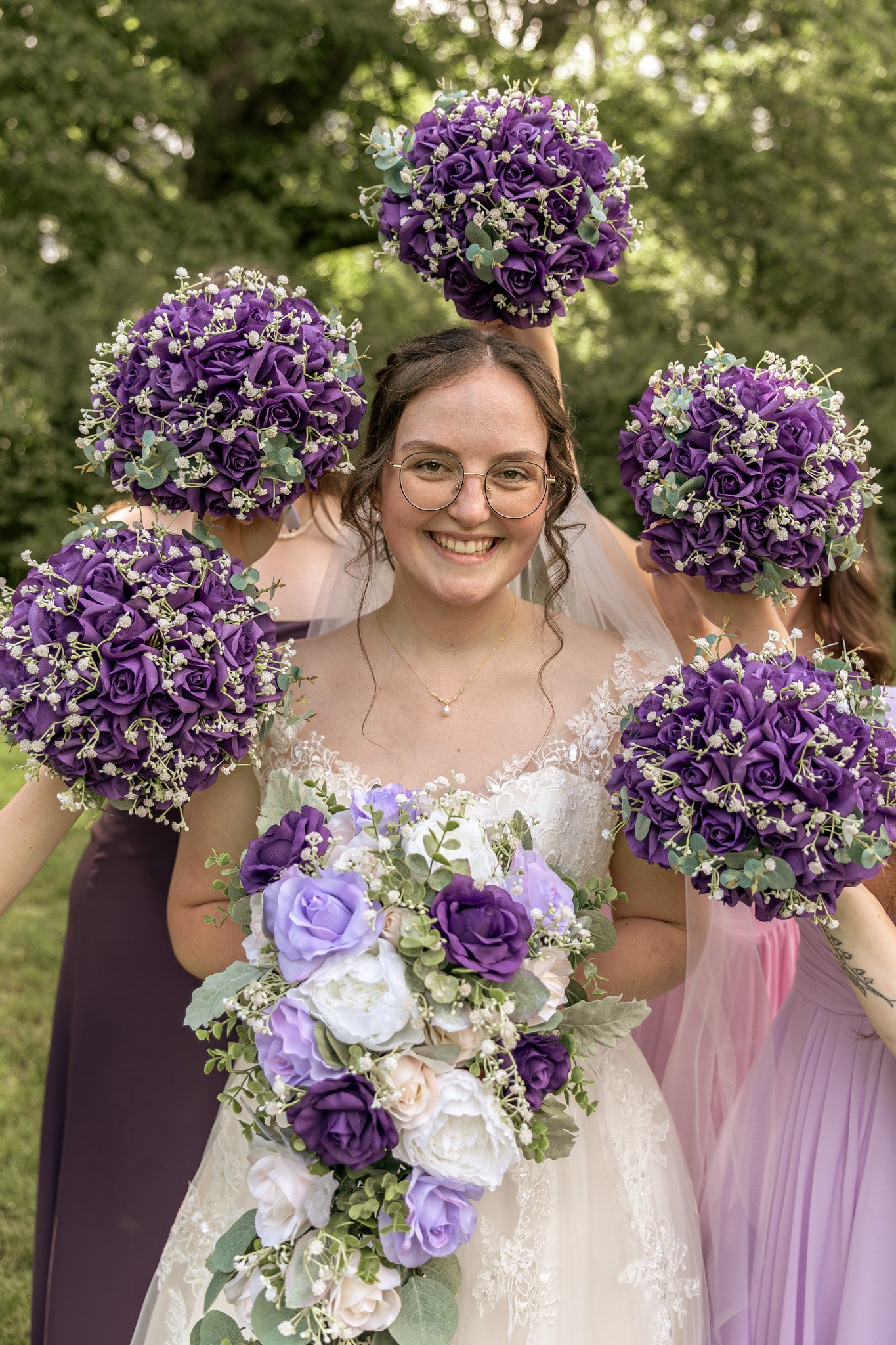 A smiling bride holding a cascading bouquet of purple, white, and lavender flowers, surrounded by bridesmaids in pink and purple dresses, outdoors with trees in the background.