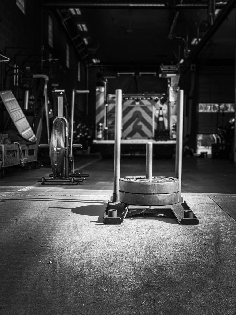 Black-and-white photo of a weighted sled loaded with plates on the floor of a fire station apparatus bay, with a fire engine and an air bike in the background.