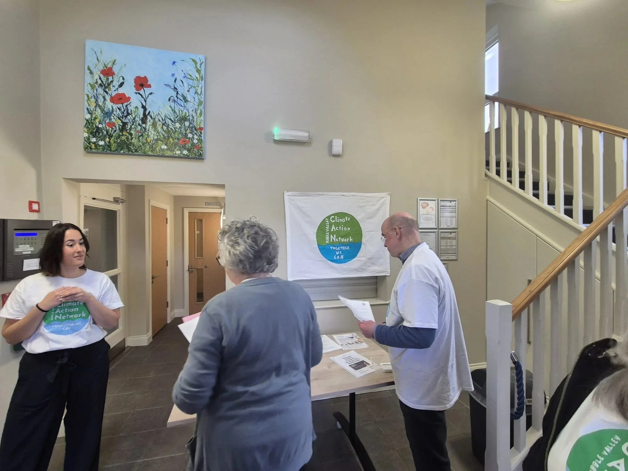 A group of people talking in a lobby area, with a woman wearing a 'Climate Action Network' t-shirt, a woman with gray hair, and a man with glasses holding papers. There is a large abstract painting on the wall, a staircase, and a table with informational materials.