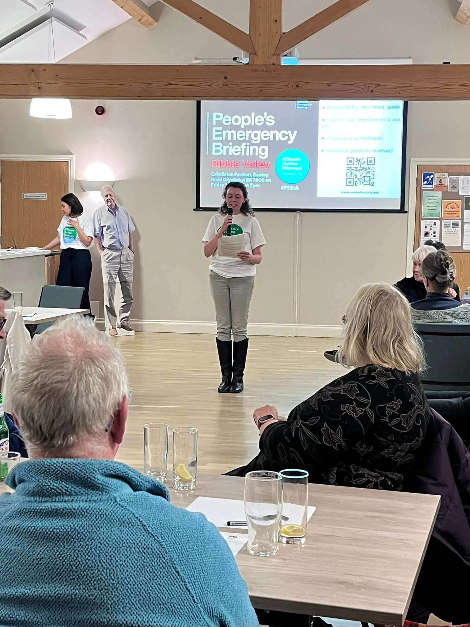 A group of people attending a presentation of a people’s emergency briefing. The presenter, a woman, is standing in front of a large screen with a microphone and notes. Others sit around tables, some with drinks and notes, listening to the presentation.