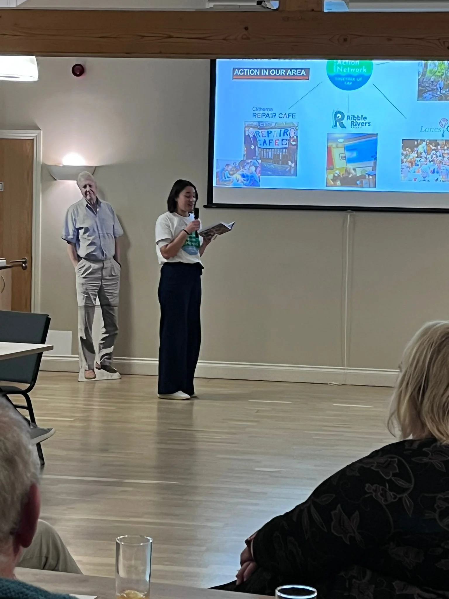 A woman giving a presentation in front of an audience, standing near a large screen displaying a slide with various images and text. An older man stands slightly behind her, near a door. Audience members are seated, and some have drinks on the table in front of them.
