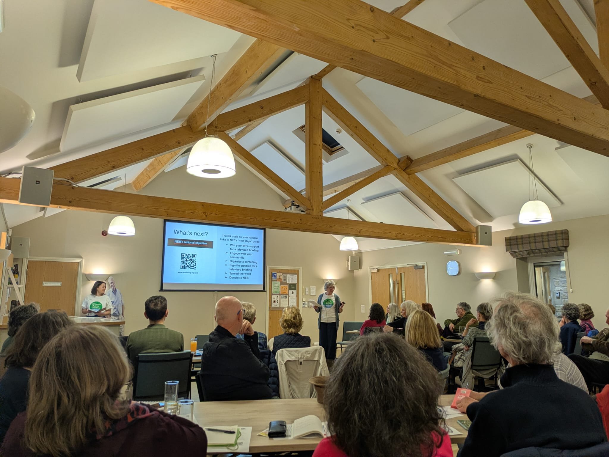 A group of people attending a presentation in a room with wooden beams on the ceiling. Two women are standing at the front, one speaking at a lectern and the other near a large screen displaying a presentation slide titled 'What's next?'. The audience is seated at tables, some taking notes or listening attentively.