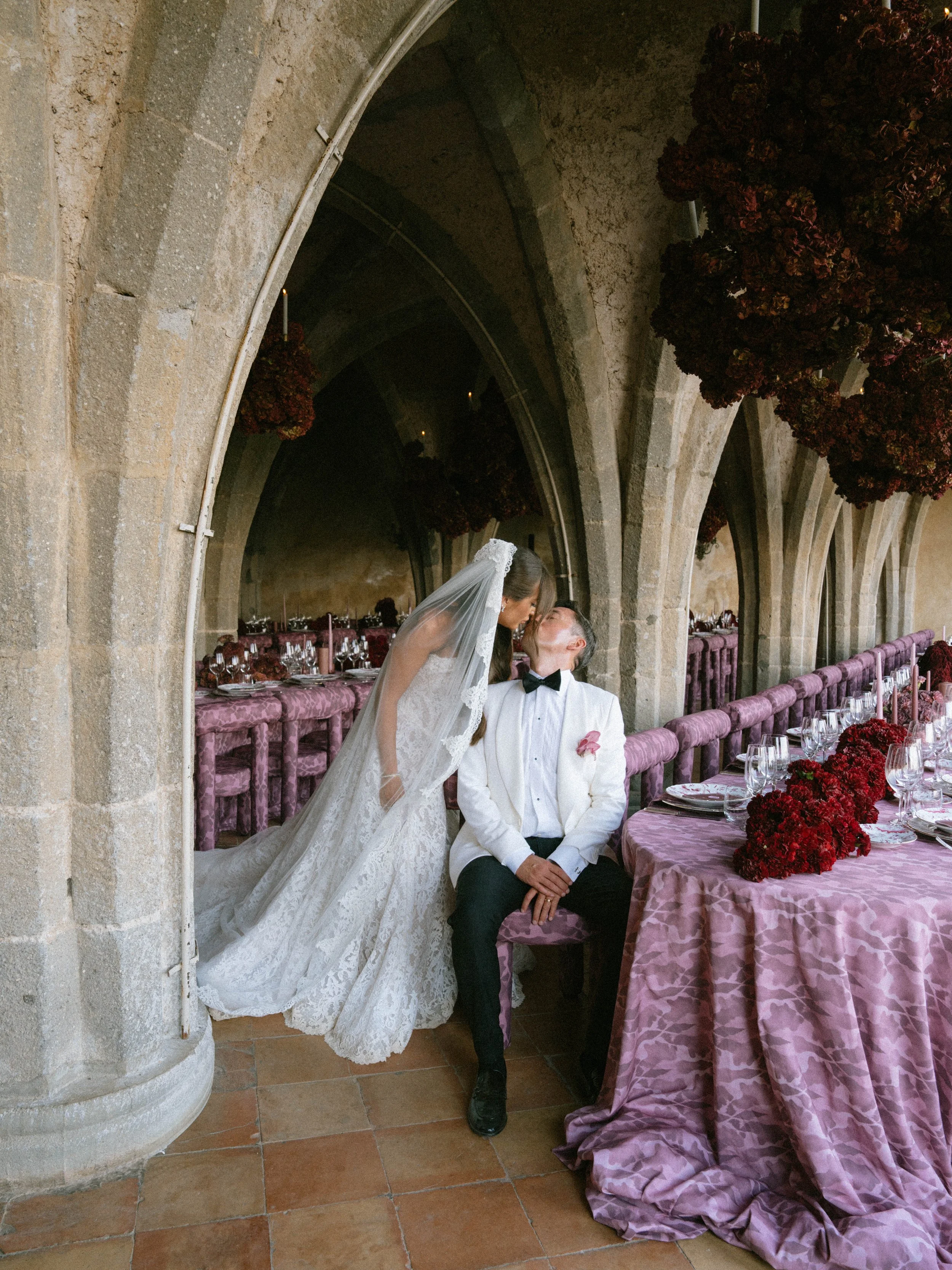 Bride and groom sitting in a stone archway at their wedding reception, with tables set with pink tablecloths and floral centerpieces. Villa Cimbrone wedding Amalfi Coast
