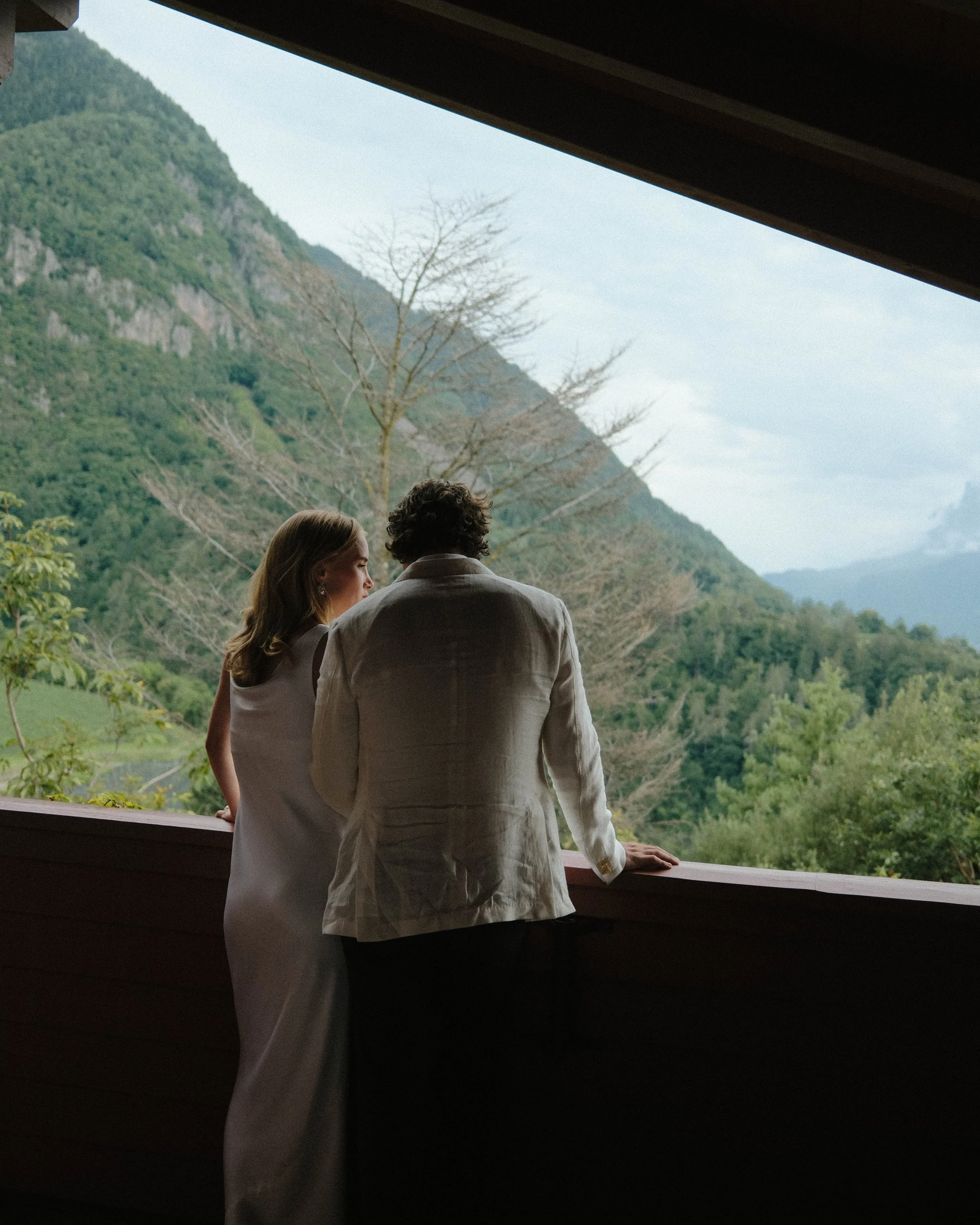 Two people, a woman and a man, standing on a balcony or terrace, looking out at a lush green mountain landscape with trees and distant mountains.