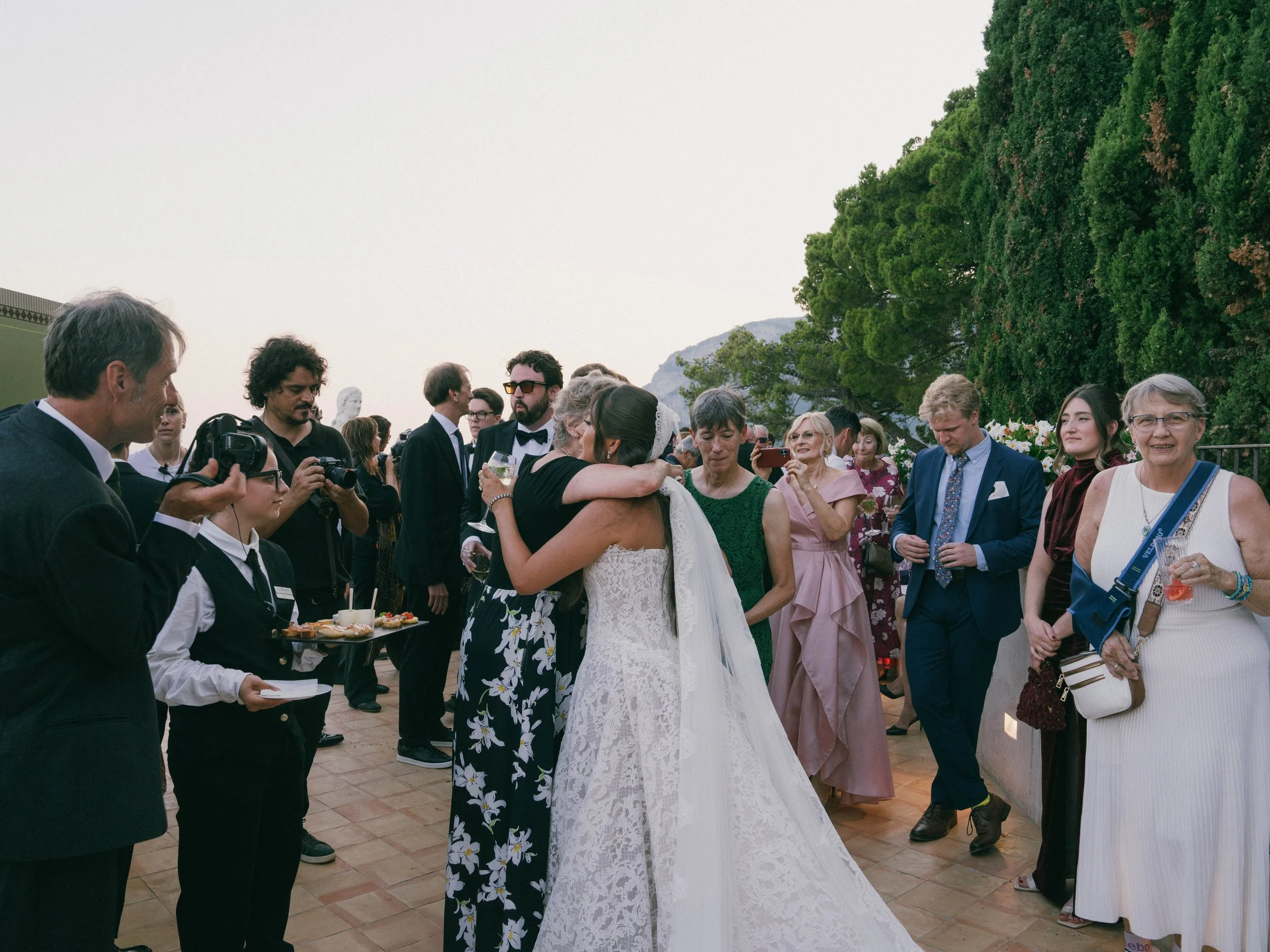 People gathered at a wedding reception outdoors, with green trees and mountains in the background, some people holding drinks, and photographers taking pictures. Villa Cimbrone wedding Amalfi Coast