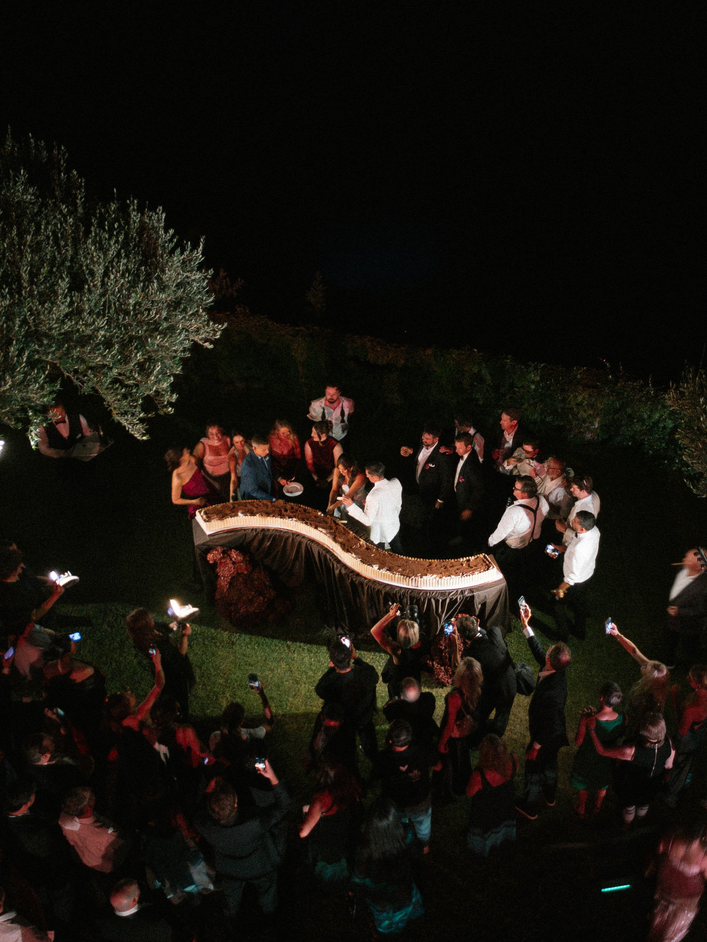 Nighttime outdoor event with a large crowd of people gathered around a long rectangular table filled with food, some people taking photos, and a priest or chef dressed in white, with trees visible in the background.