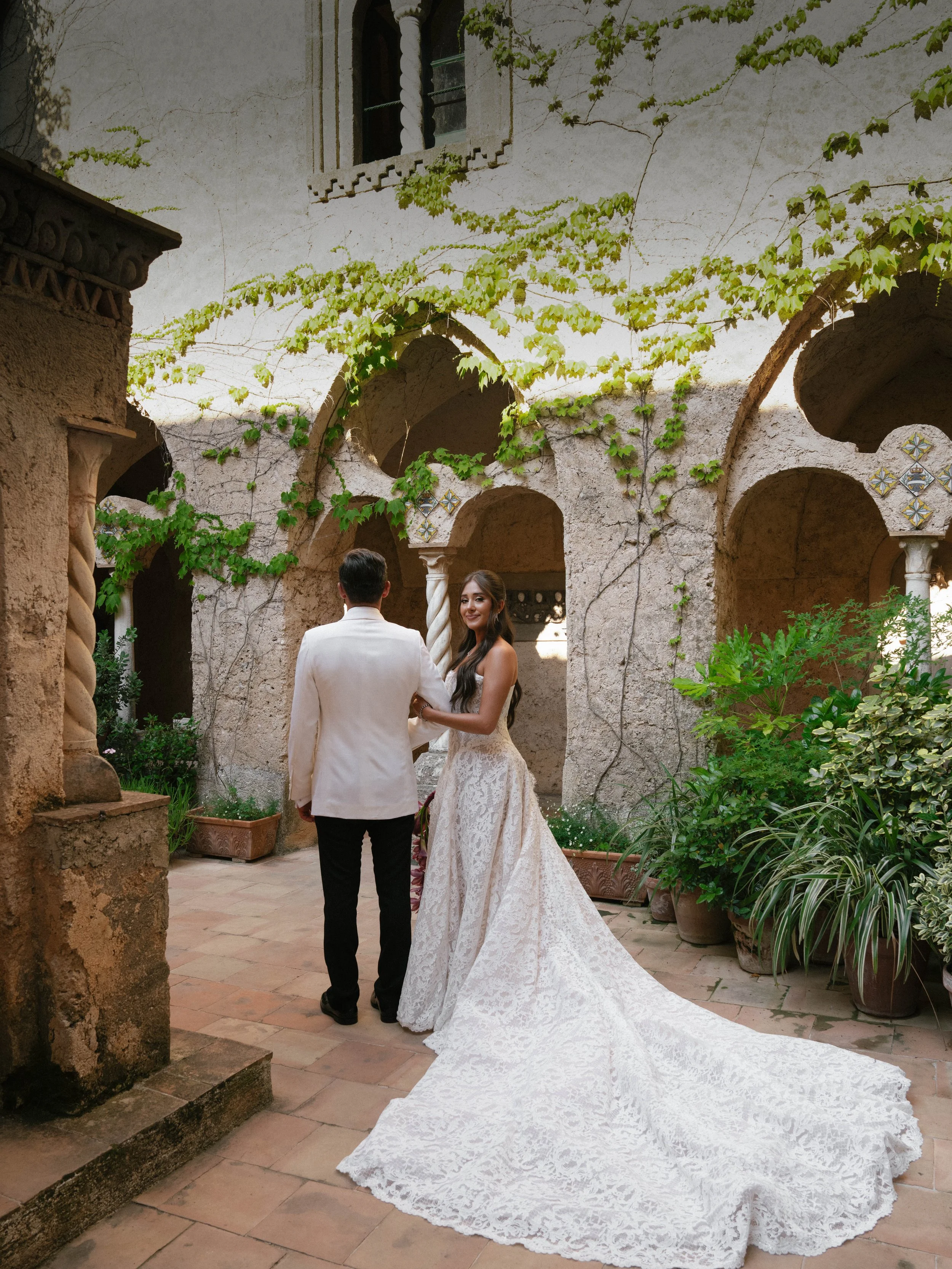 A bride and groom are holding hands in a courtyard surrounded by ivy-covered walls and potted plants, with the bride wearing a long lace wedding gown with a train.