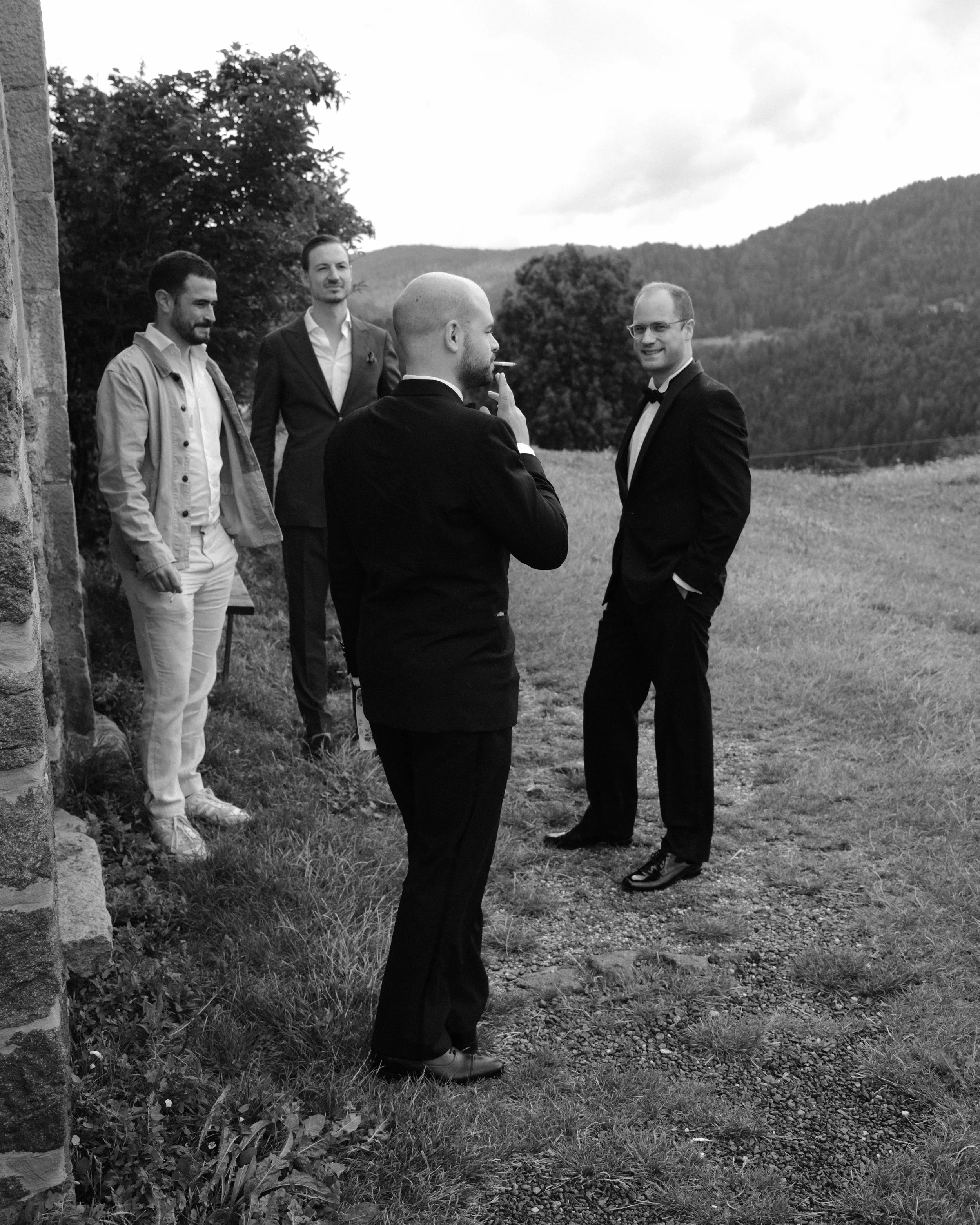 Four men in suits outdoors, one smoking a cigarette, with hills and trees in the background, black and white photo.