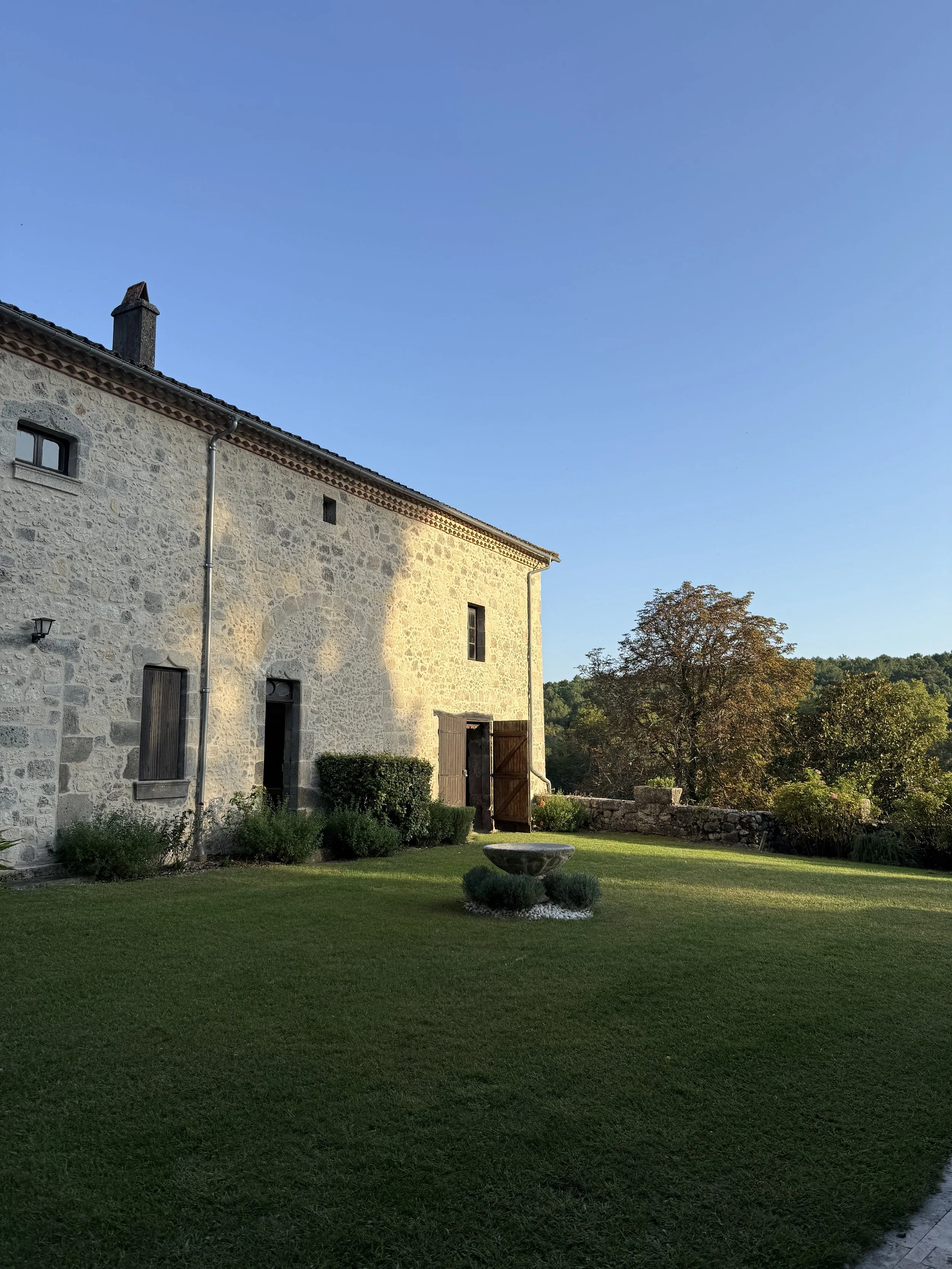 Stone house with open wooden door, green lawn, small bushes, and trees in the background under a clear blue sky.