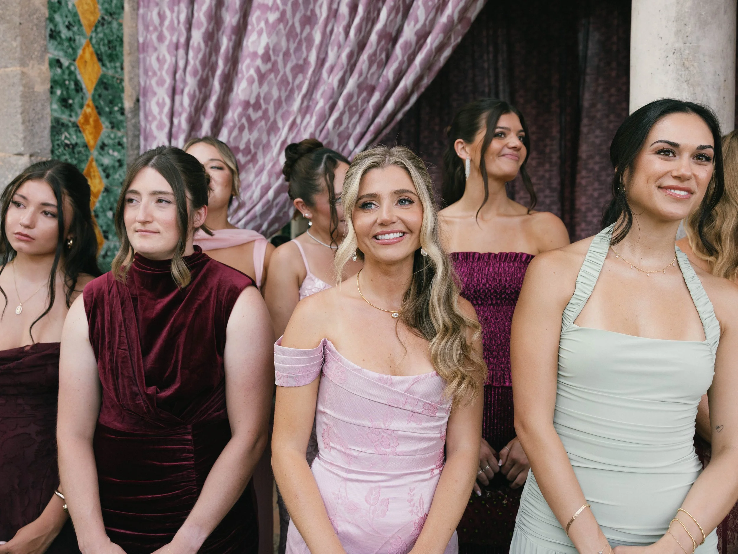 Group of women dressed in evening gowns at a formal event, standing in front of a decorated backdrop with purple curtains and green and yellow patterned fabric.