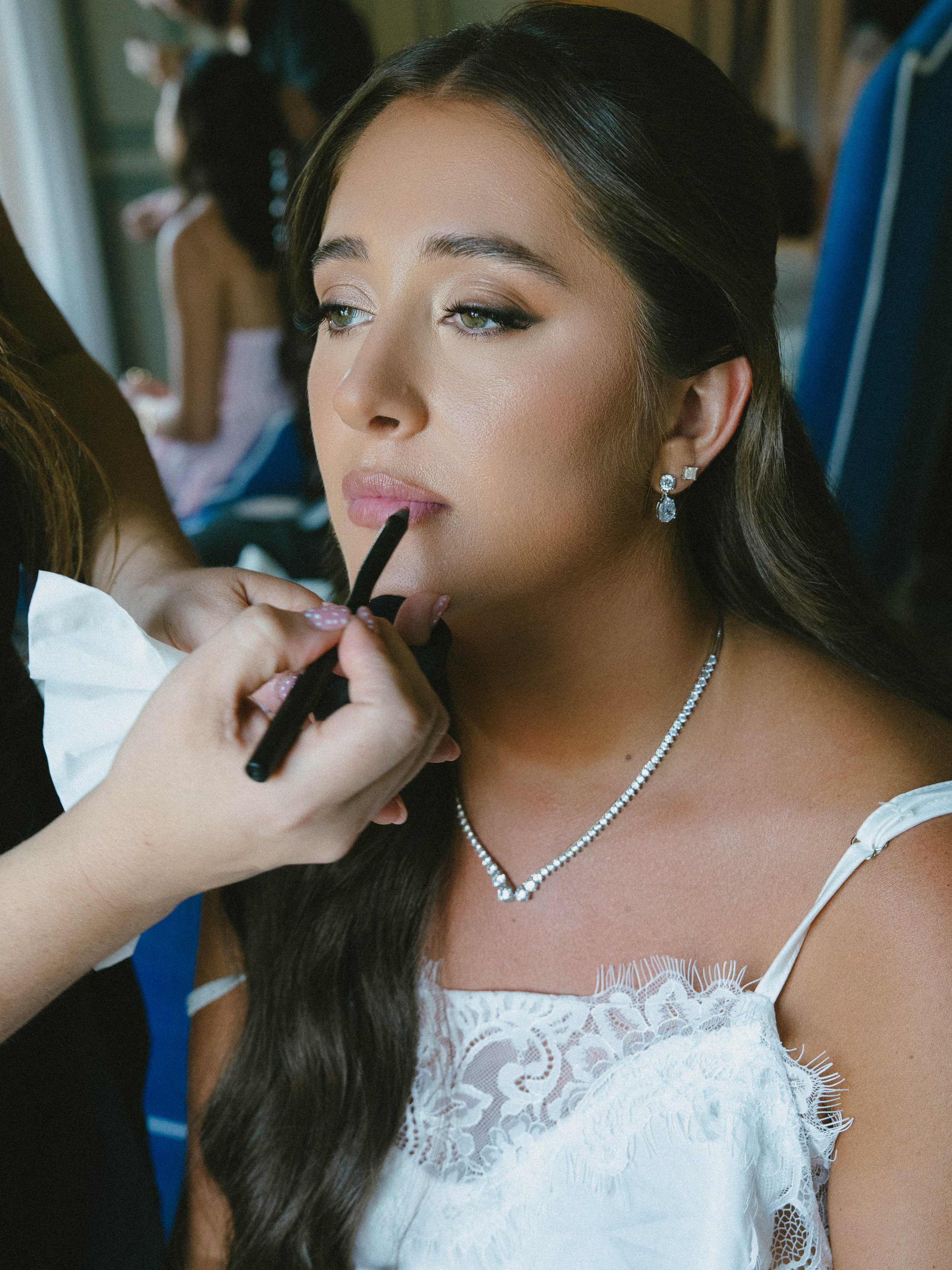 A woman with long dark hair and light skin, wearing jewelry, is getting her makeup done by a makeup artist applying lipstick in what appears to be a special occasion setting. She is dressed in white lace attire and has a serene expression.