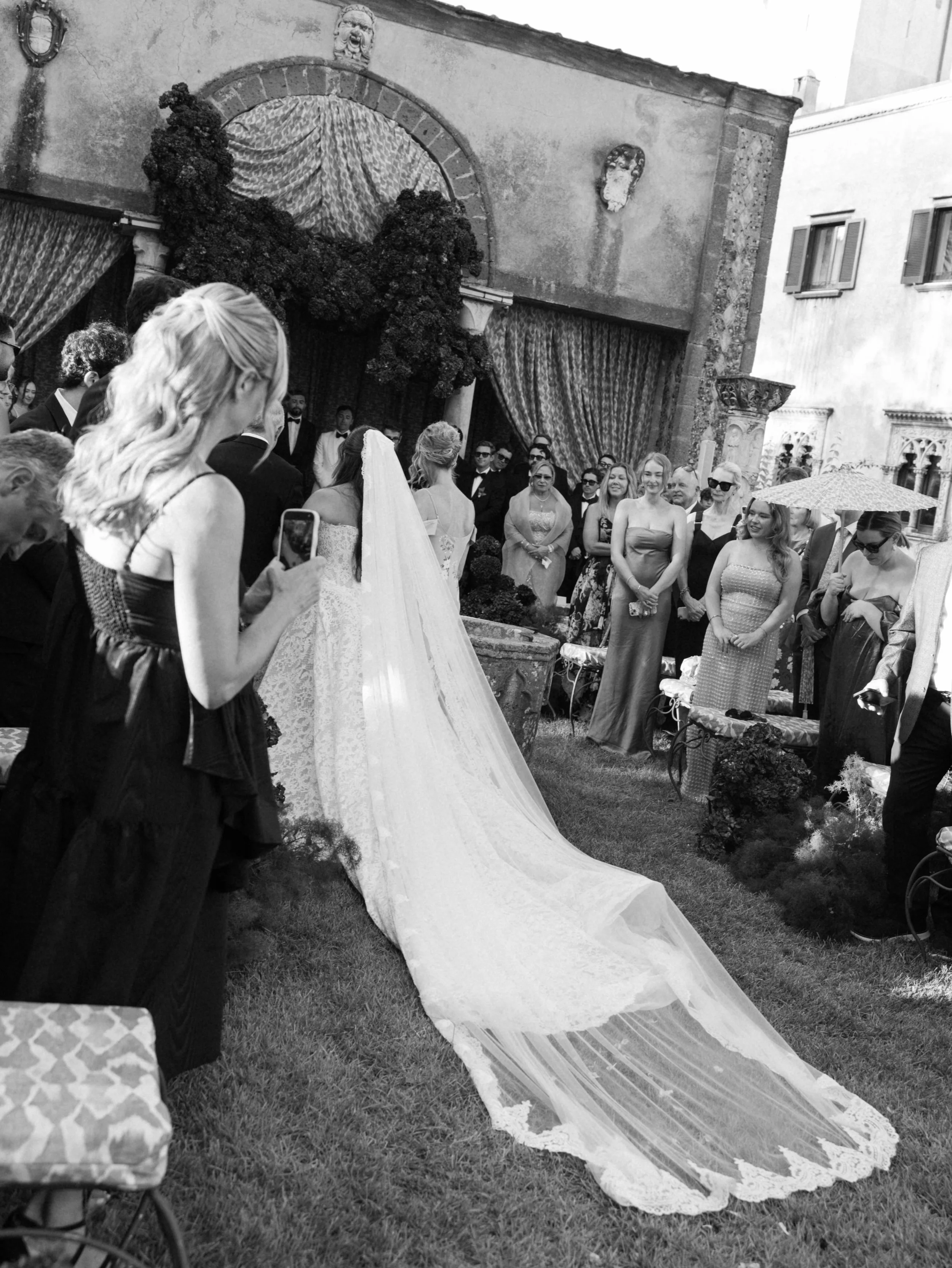 People gathered outdoors for a wedding ceremony, with a bride in a white gown and veil, some guests holding umbrellas, and decorative drapes and masks on a building backdrop.