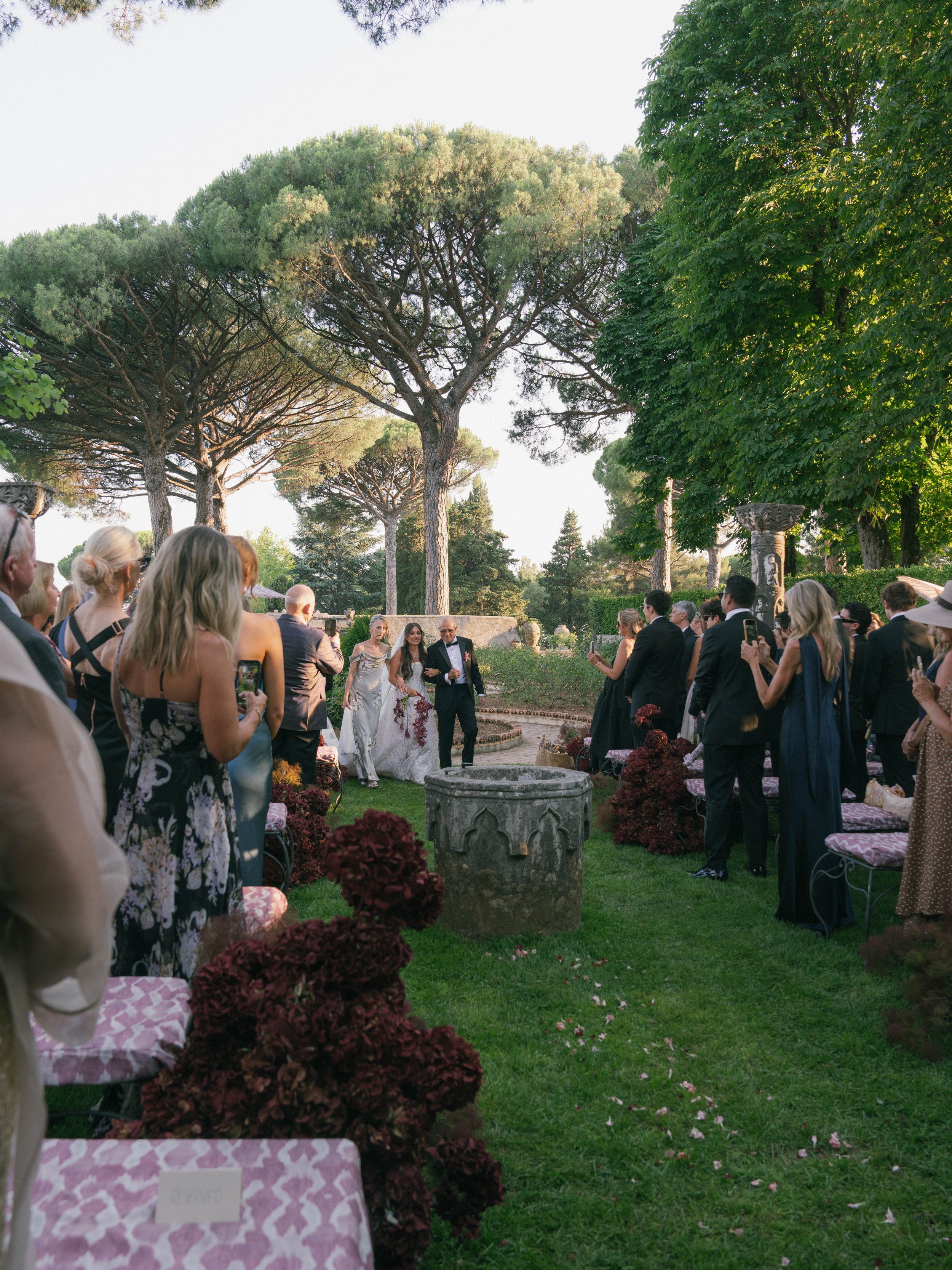 A wedding ceremony outdoors with guests seated on pink patterned chairs, greenery and tall trees in the background, and a couple walking together in the center.Villa Cimbrone wedding Amalfi Coast