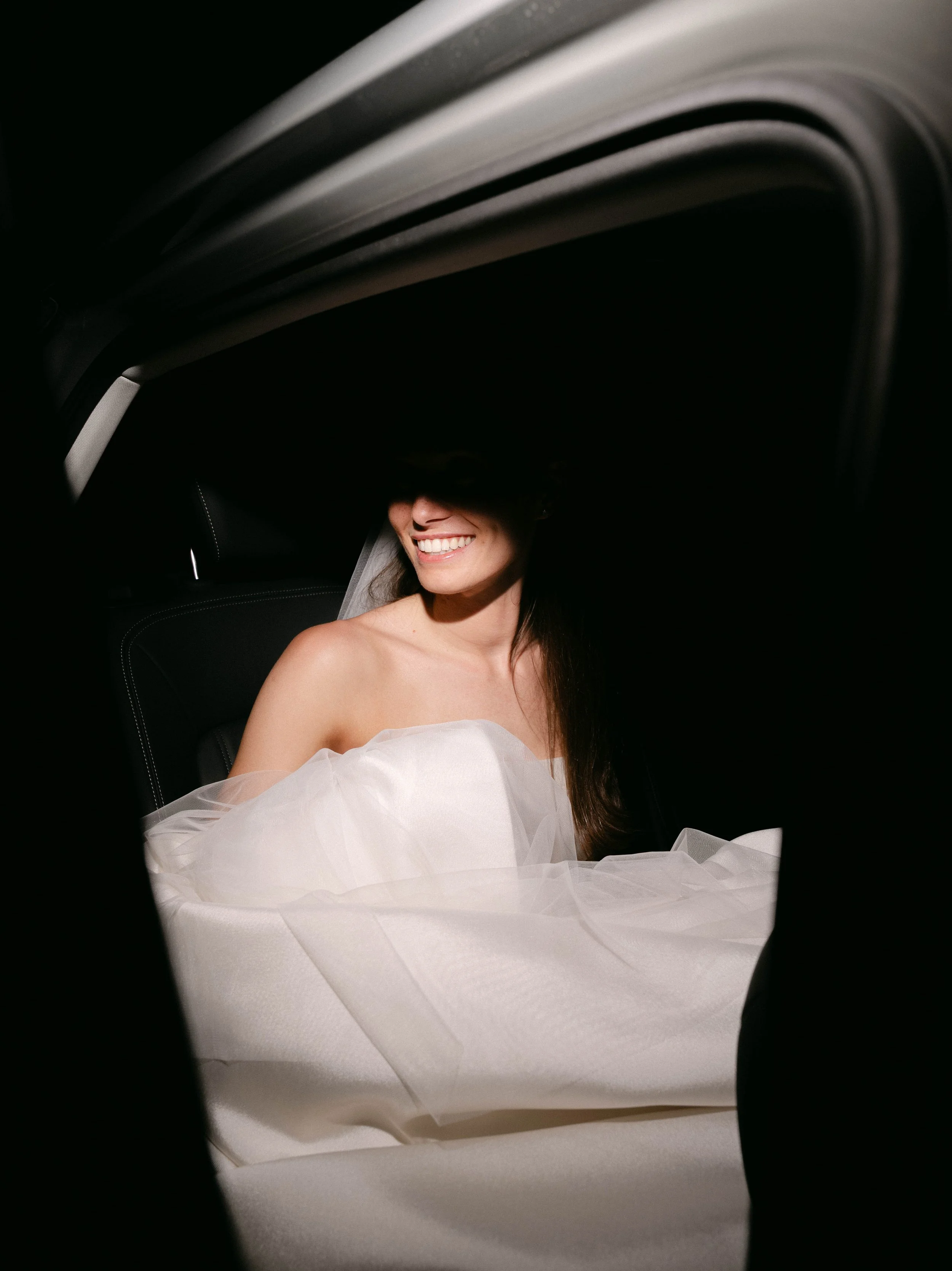 A bride in a bridal dress sitting in a vehicle, seen through a small opening or window with a black border.