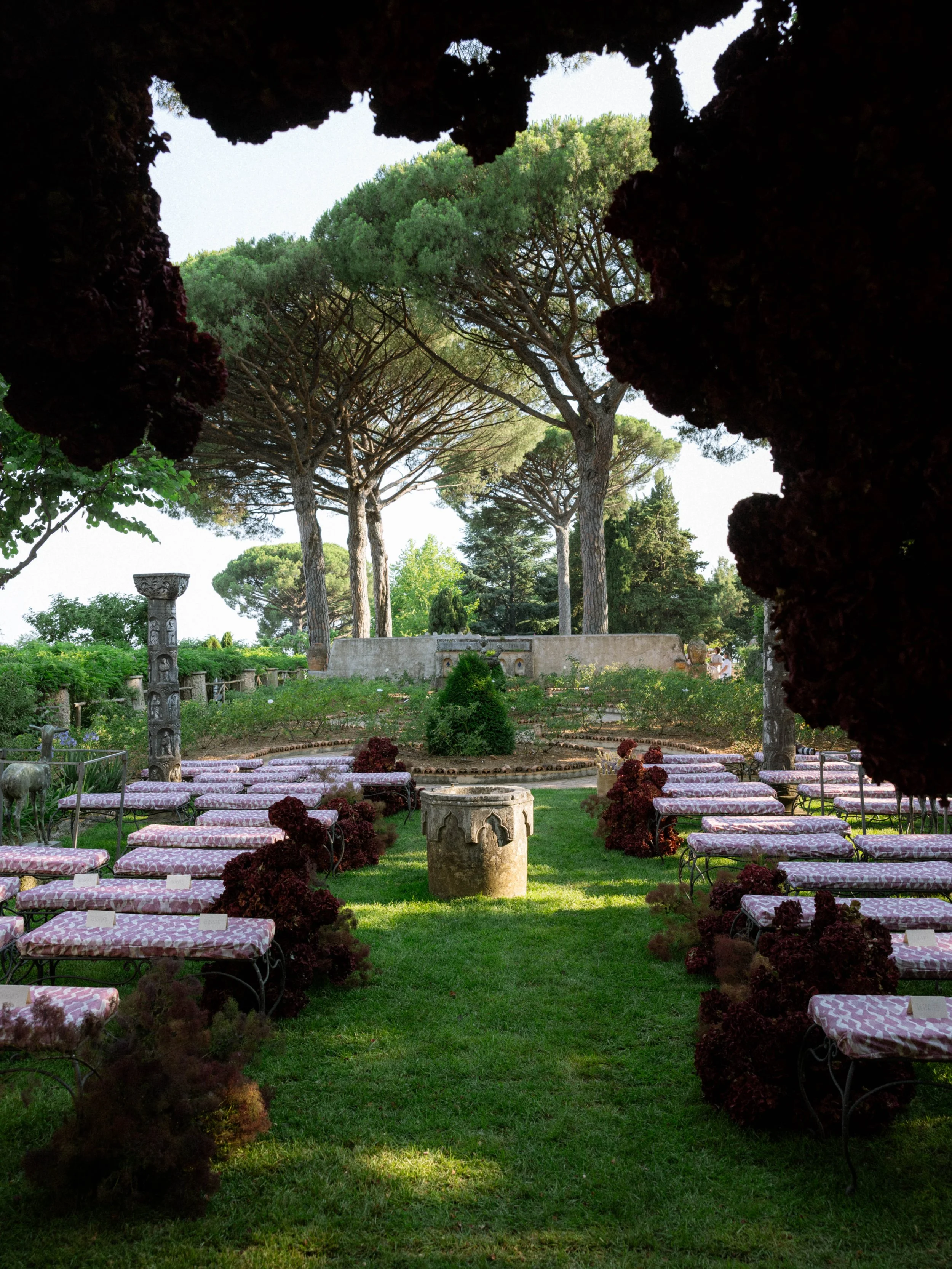A garden scene viewed through a dark opening, with pink and white patterned benches arranged around a small central area, decorated with purple flowers, with large trees and greenery in the background.