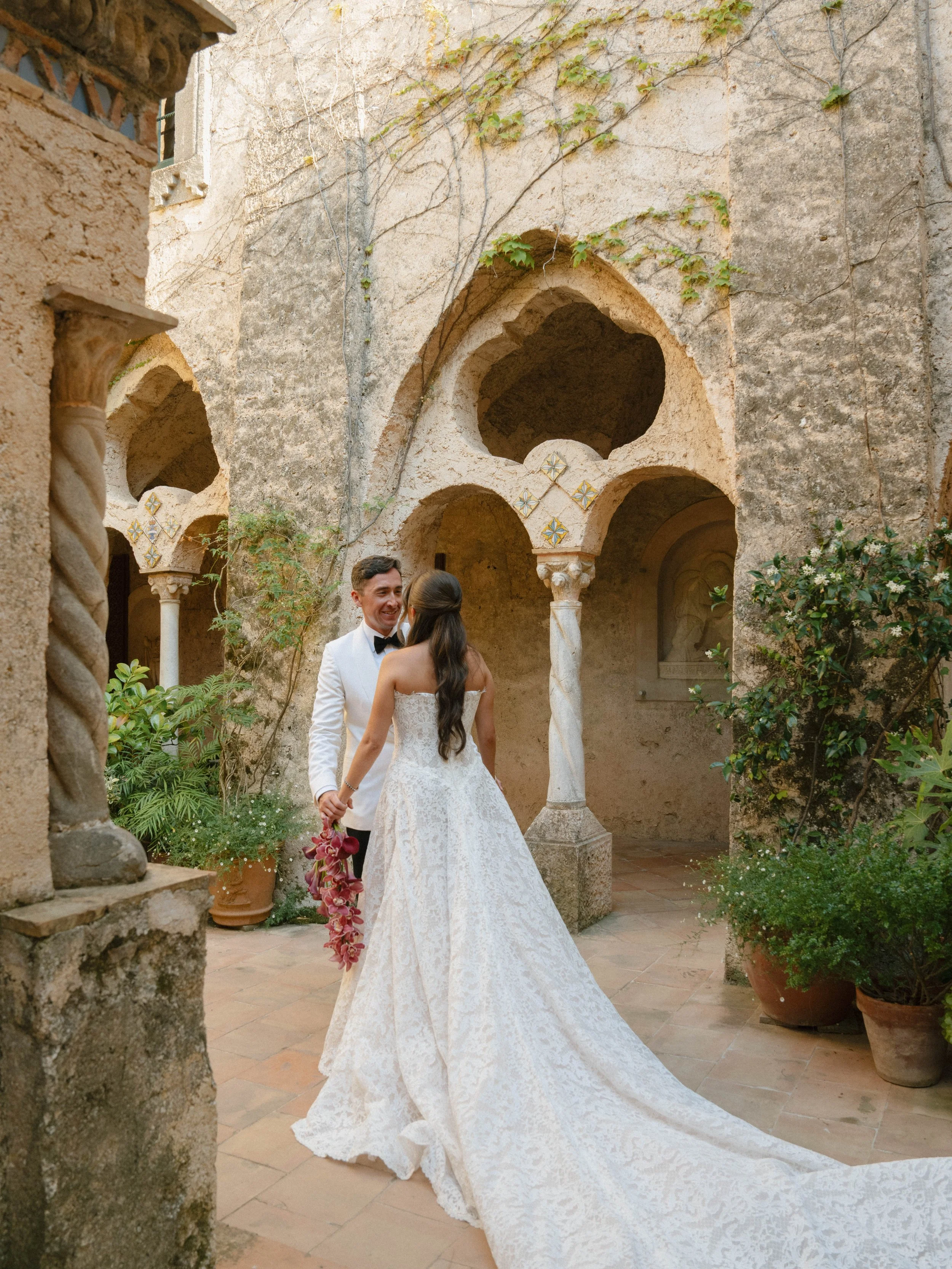 A bride and groom hold hands in a courtyard with stone arches, planters, and greenery, during a wedding celebration.