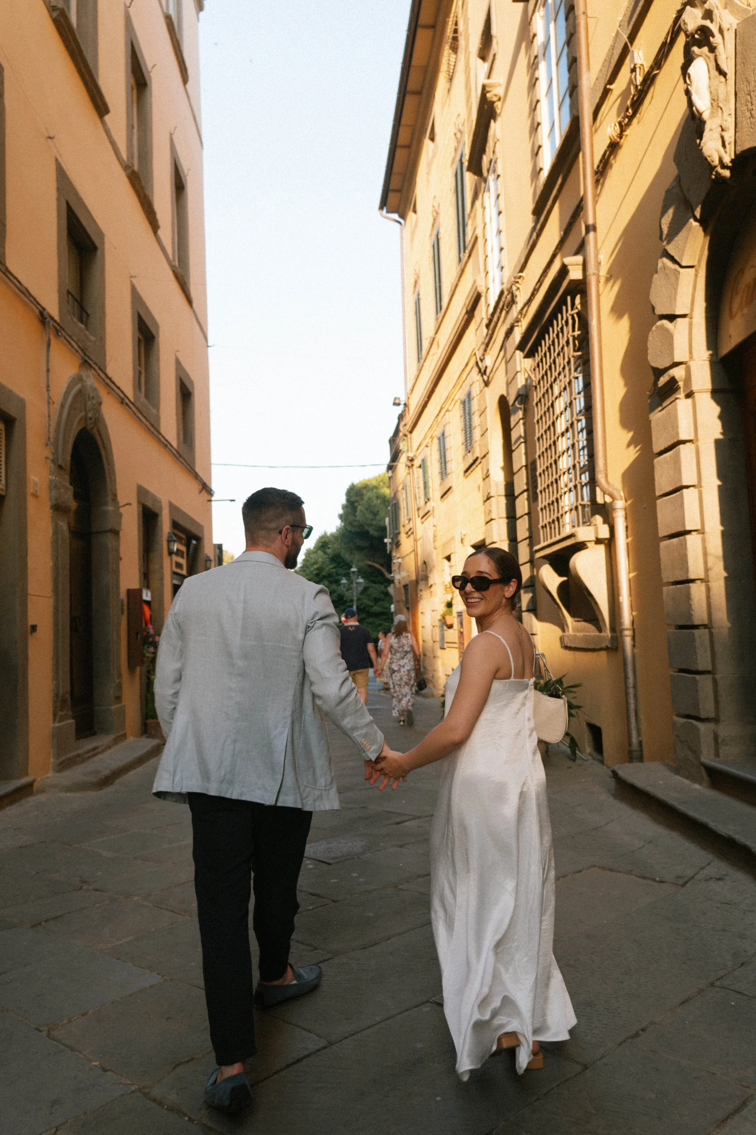 A couple holding hands and walking down a narrow street in a European city, with the woman smiling at the camera, both dressed in stylish clothes during daylight.