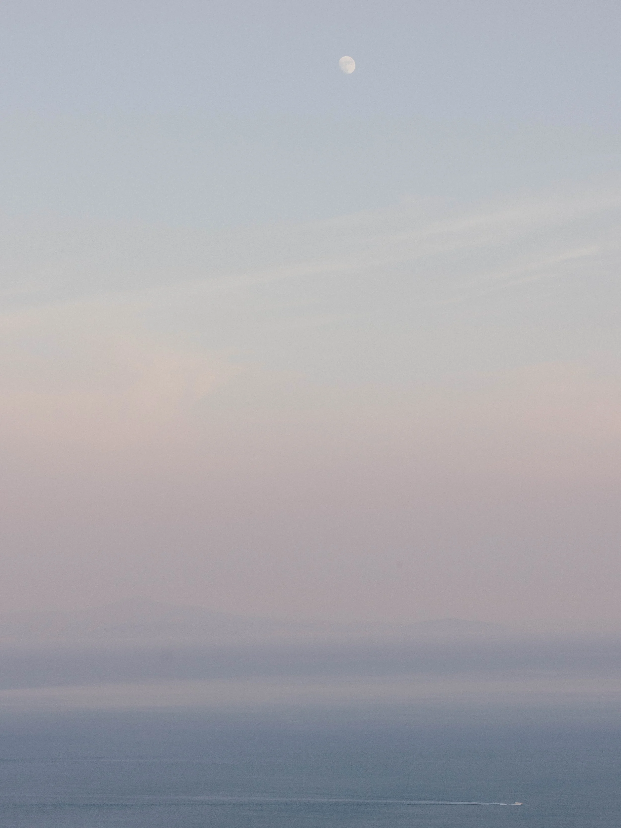 A view of the sky with the moon visible in the upper center, and a calm ocean with a small boat passing through the water in the lower right.