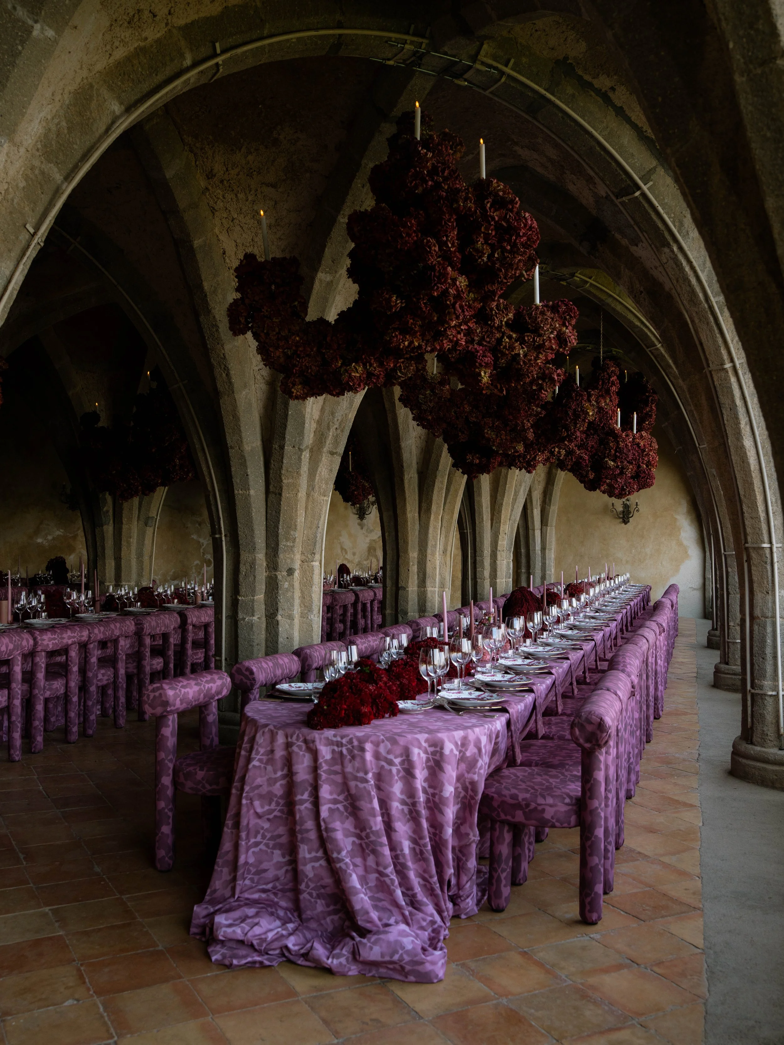 A long dining table set with glassware, plates, and red floral centerpieces in a stone vaulted room with large floral arrangements hanging from the ceiling. Villa Cimbrone