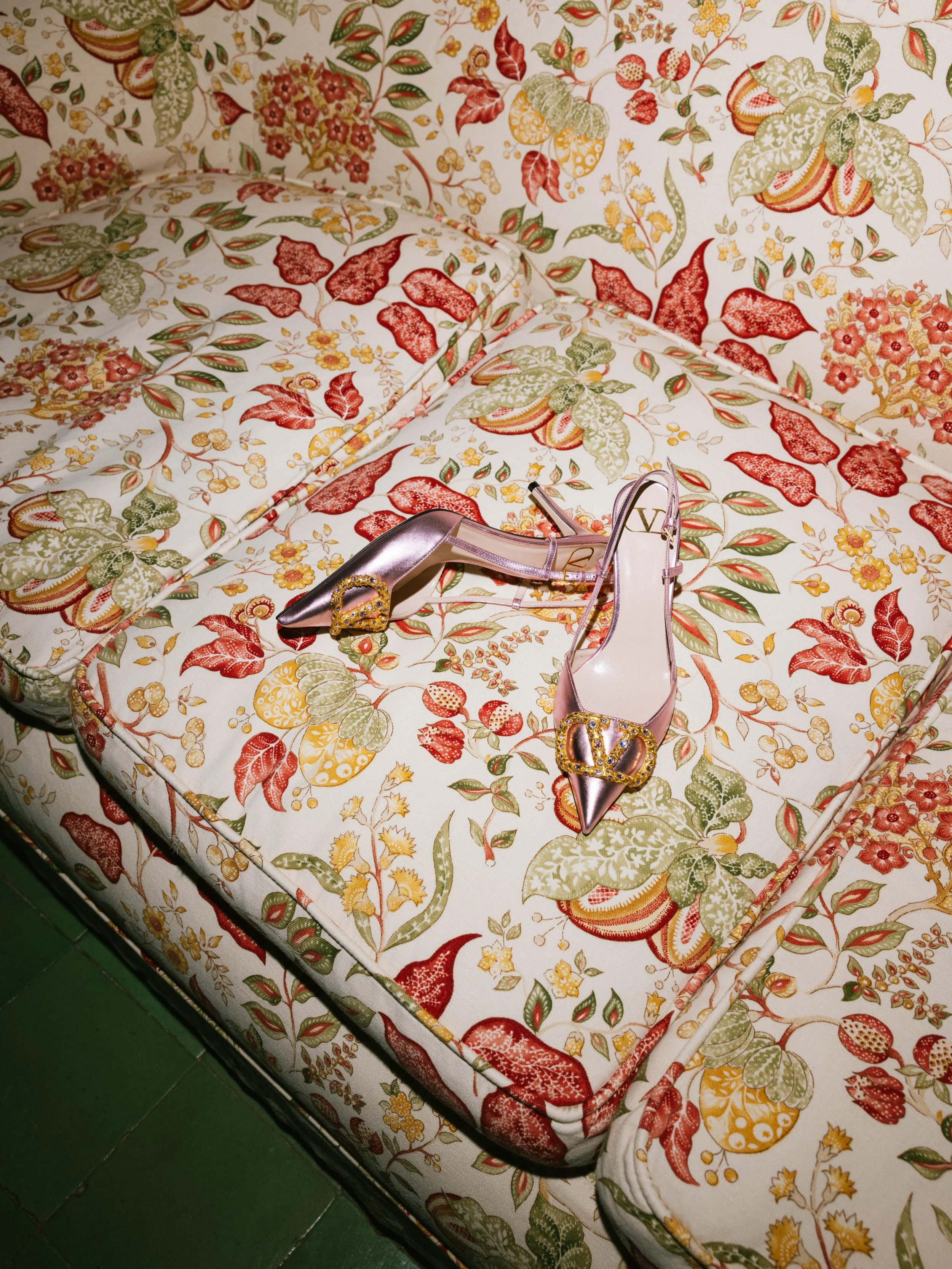 A floral patterned bedspread with a pair of shiny pink high-heeled shoes with jeweled buckles resting on it.