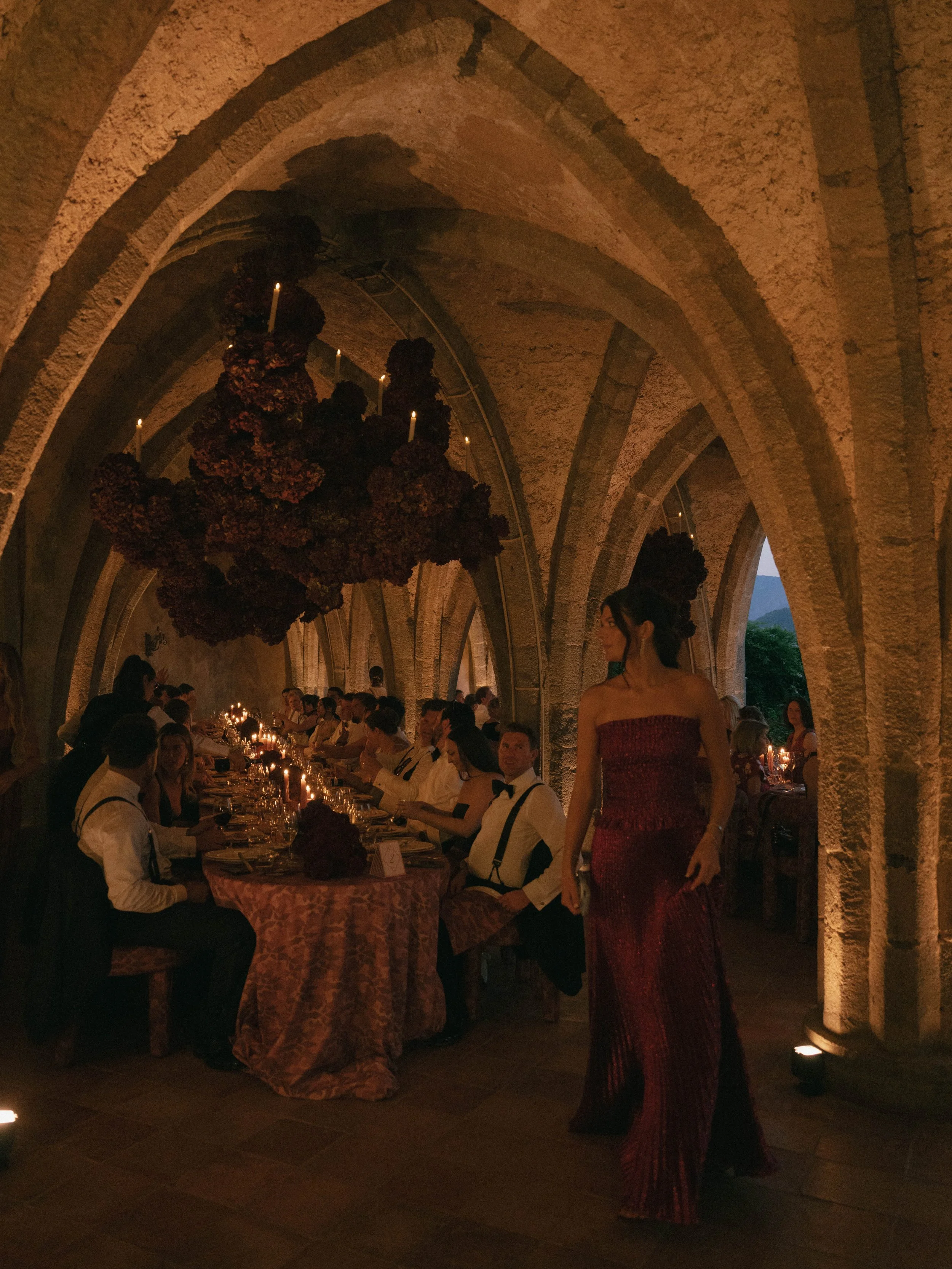 A woman in a red, strapless, sparkly gown standing near a long dining table filled with people, candles, and floral centerpieces inside a stone archway at a formal event.