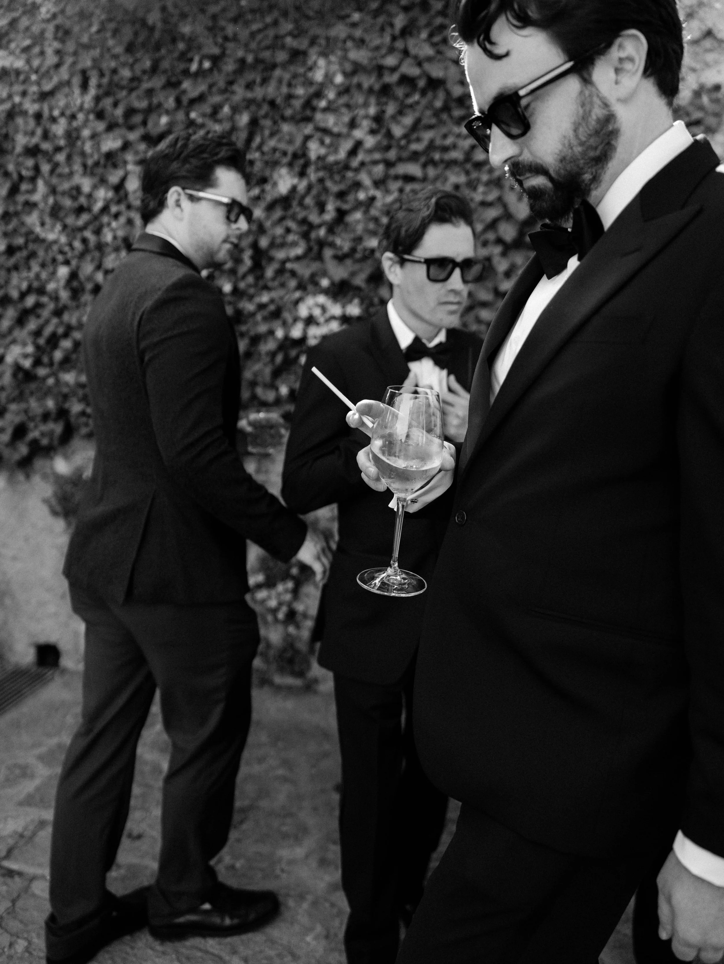 Four men in tuxedos and sunglasses standing outdoors, one holding a glass of wine and a cigarette, with a leafy hedge in the background.