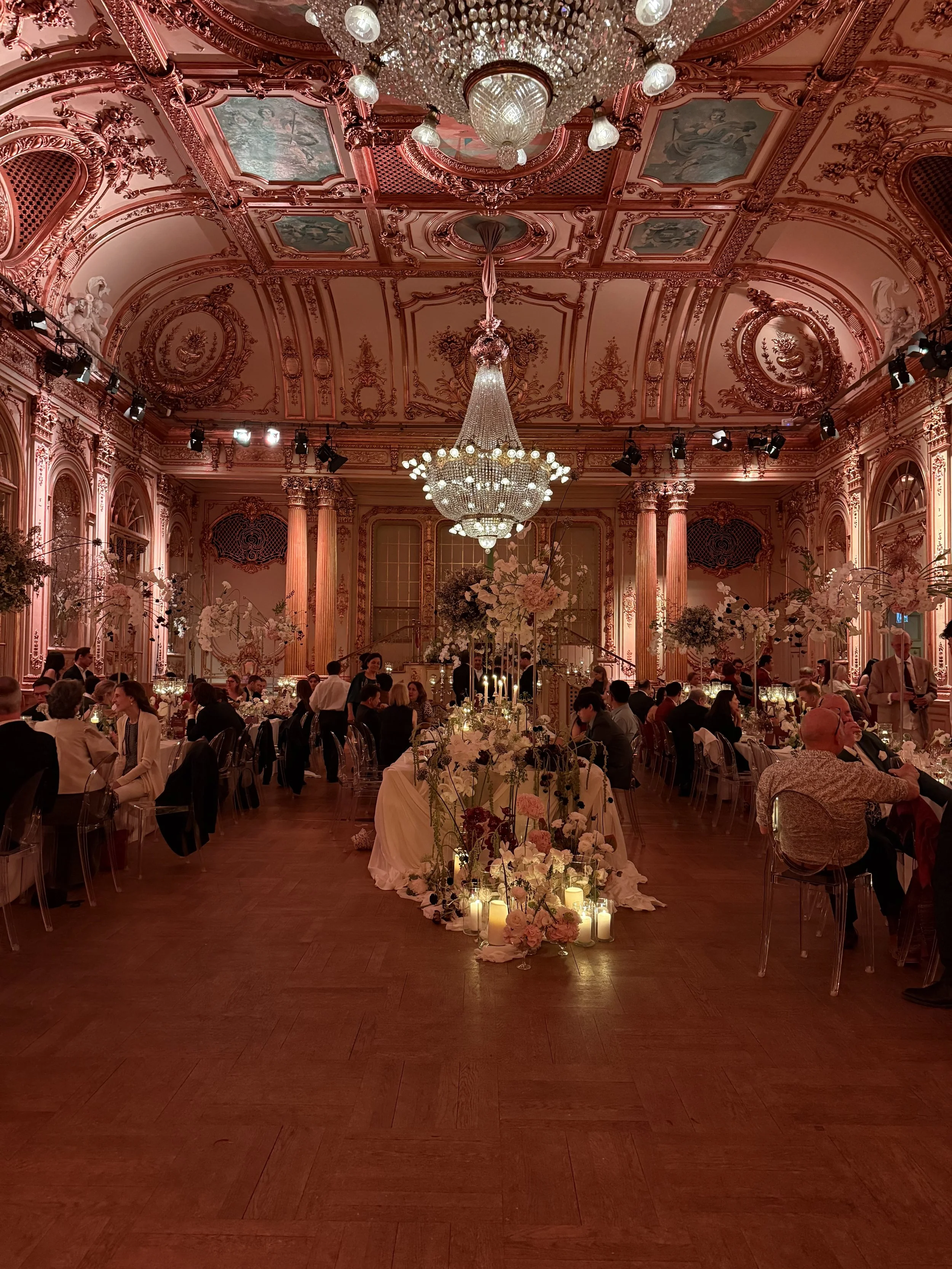 Elegant ballroom decorated for a formal event, featuring a grand chandelier, ornate pink and gold walls, and long banquet tables adorned with floral centerpieces and candles.