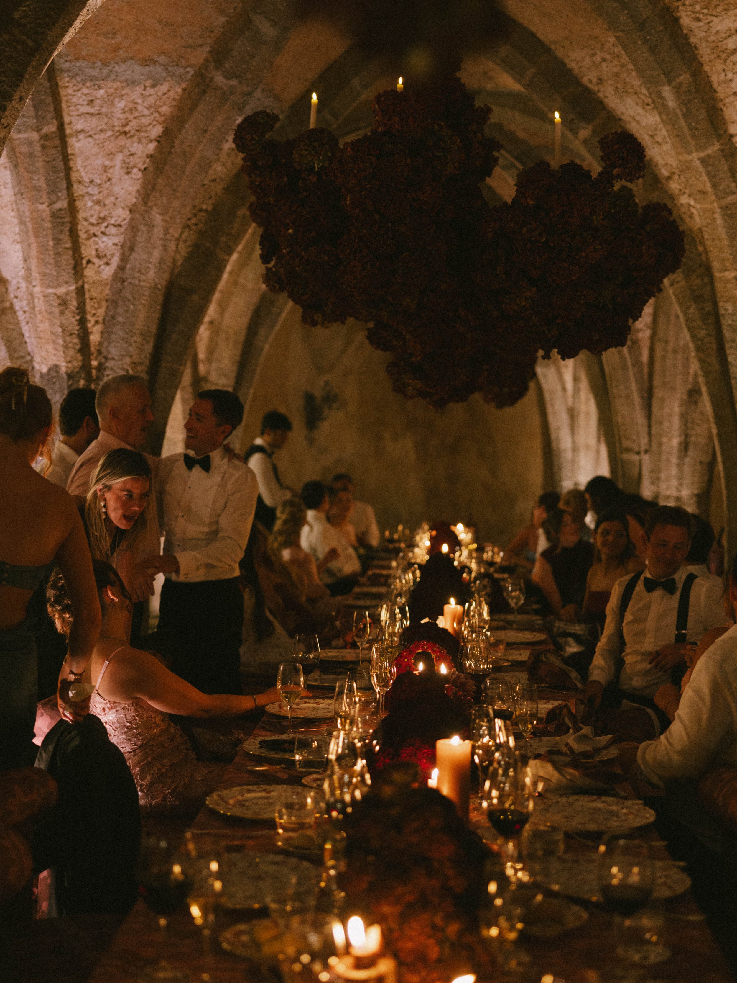 People gathered around a long dining table with candles, in a dimly lit stone vaulted room. A dark chandelier hangs from the ceiling, decorated with flowers or organic material, and candles are also on the table.