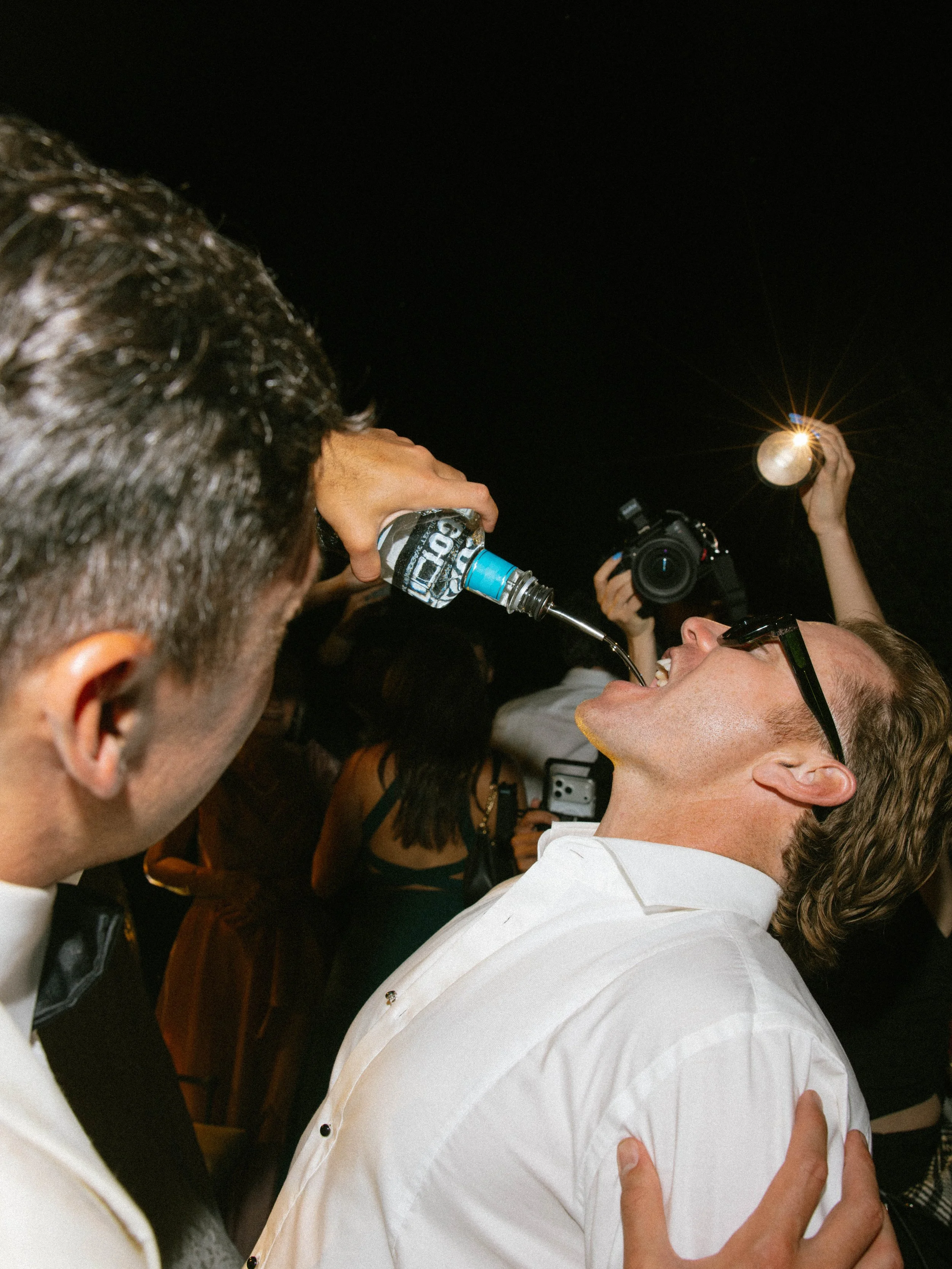 Several people celebrating at night, one person holding a flashlight, another person holding a water bottle, and a third person under the flashlight's light, all smiling and wearing formal attire.