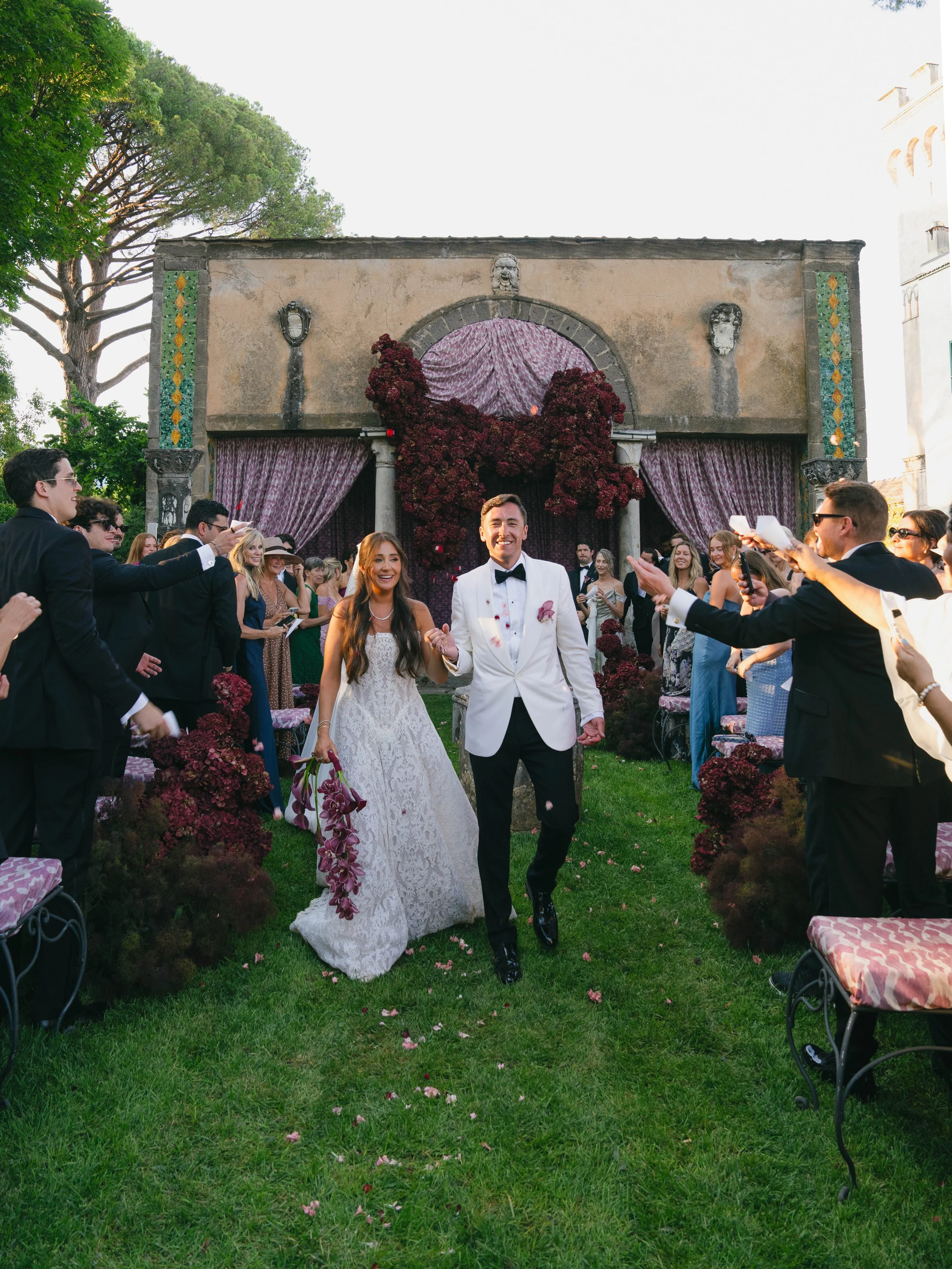 A bride and groom walking down the aisle at an outdoor wedding ceremony with guests on both sides. The bride is wearing a white lace wedding dress and holding a bouquet, and the groom is wearing a white tuxedo jacket with black pants and a black bow 