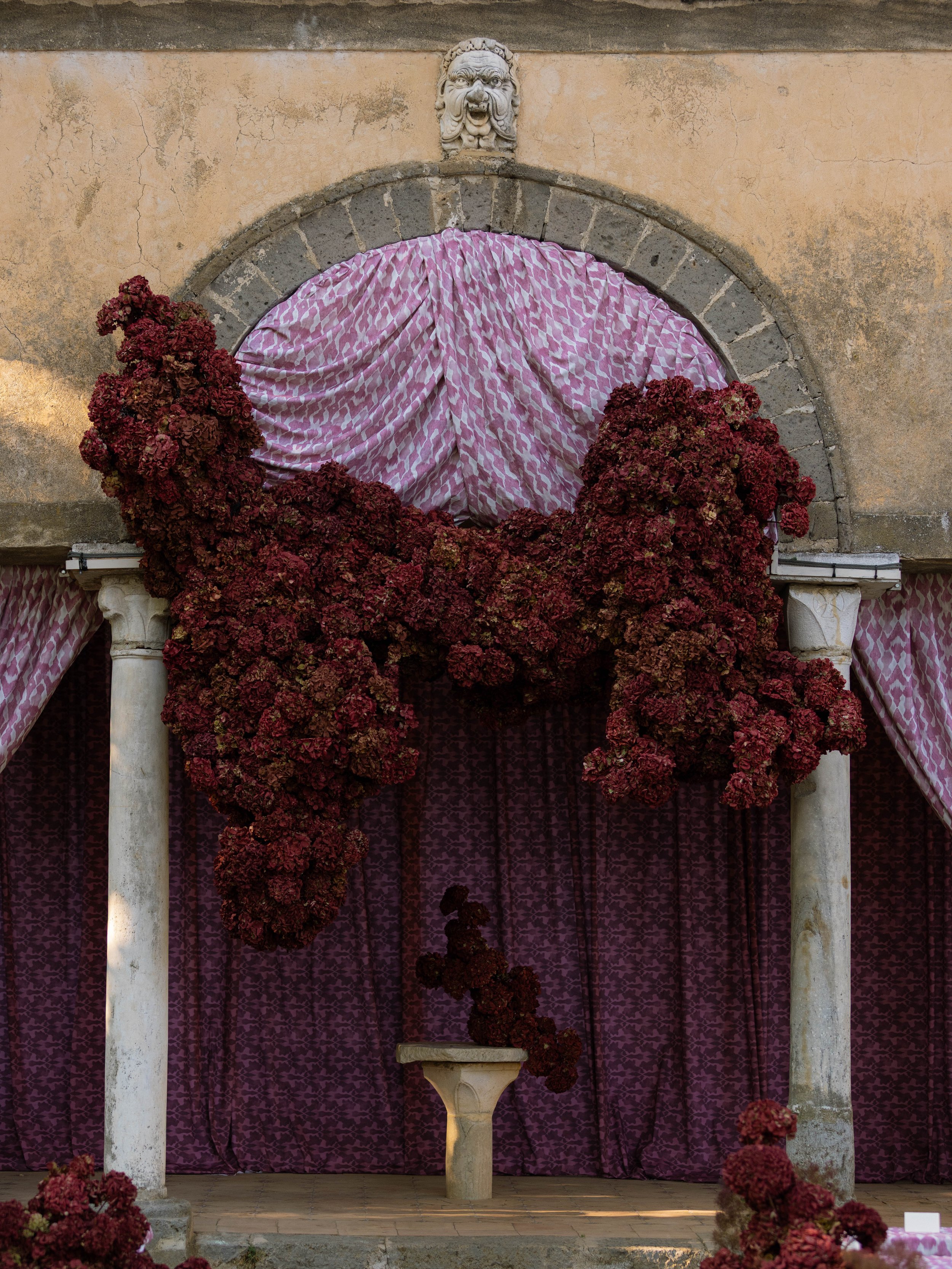 A decorative outdoor wall with an arched opening draped with pink and white patterned fabric. Two white columns support the arch, and a lion head sculpture decorates the top of the wall. Dense red flowers surround the base of the arch, and purple cur