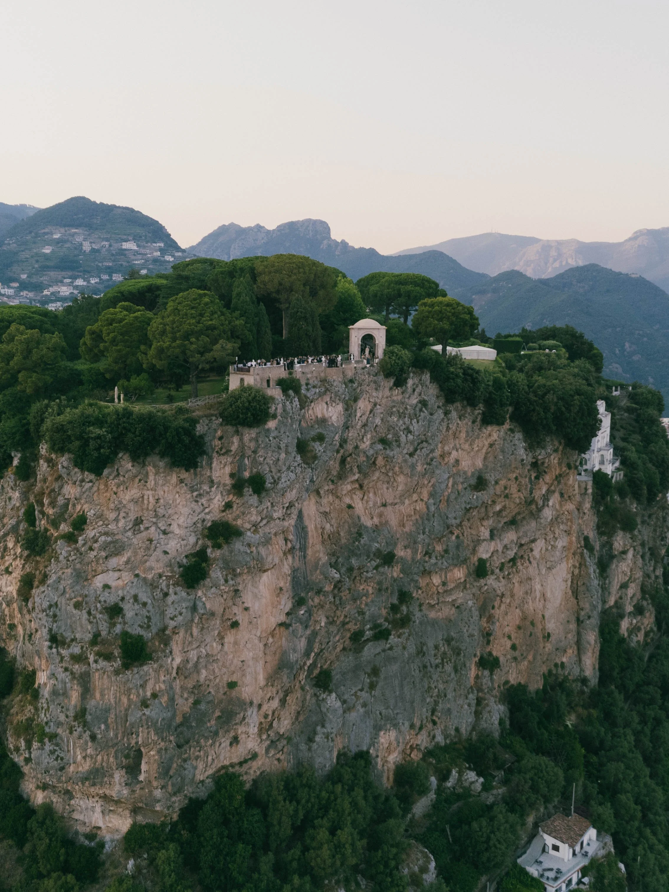 A castle-like structure with an arched entrance is perched on a rocky hillside, surrounded by lush green trees, with mountains in the background and a house at the very bottom of the hill.  Villa Cimbrone wedding Amalfi Coast Ravello Infinity Terrace