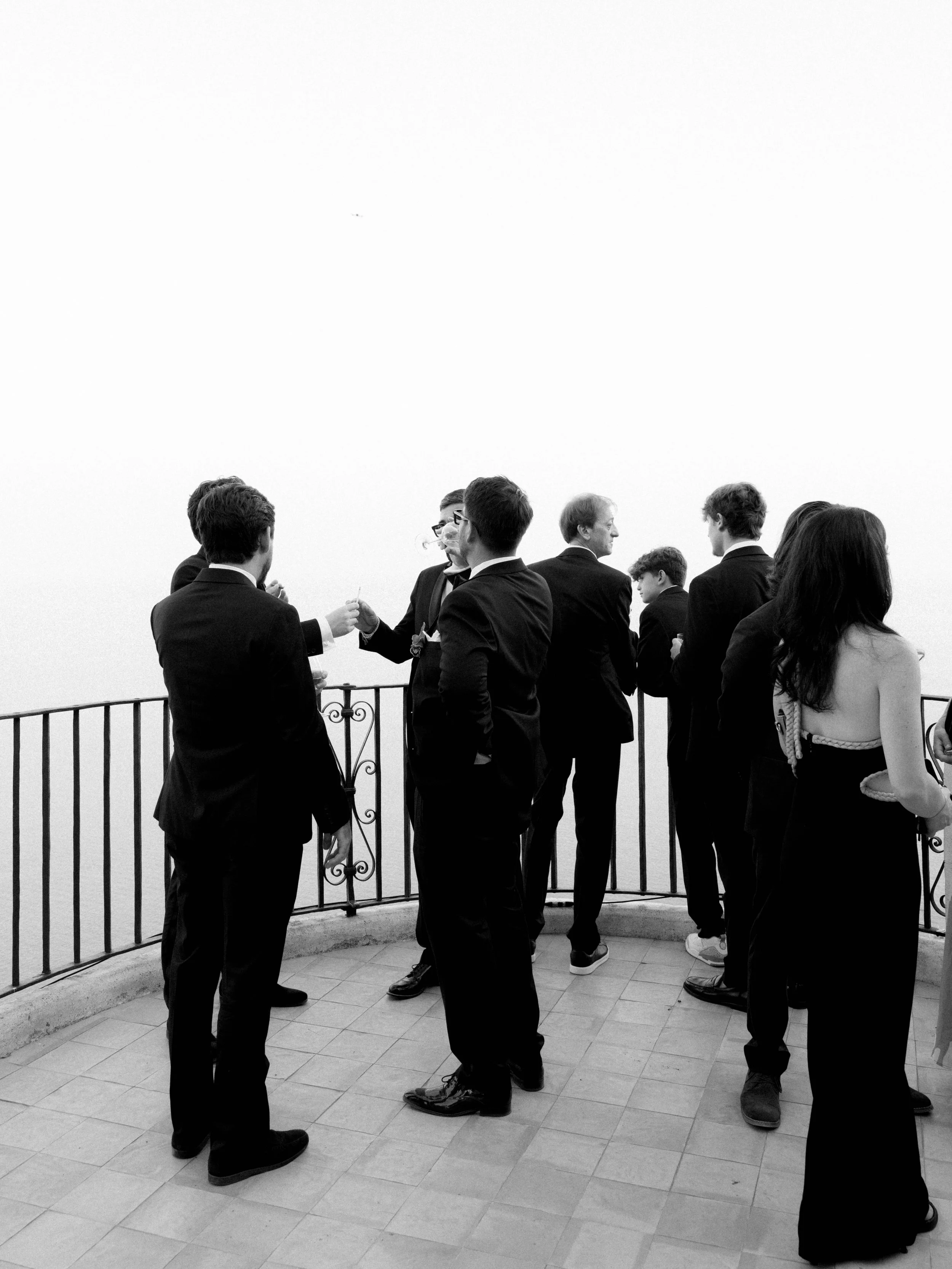 Black and white photo of a group of people in formal attire gathering outdoors near a railing, engaging in conversation.