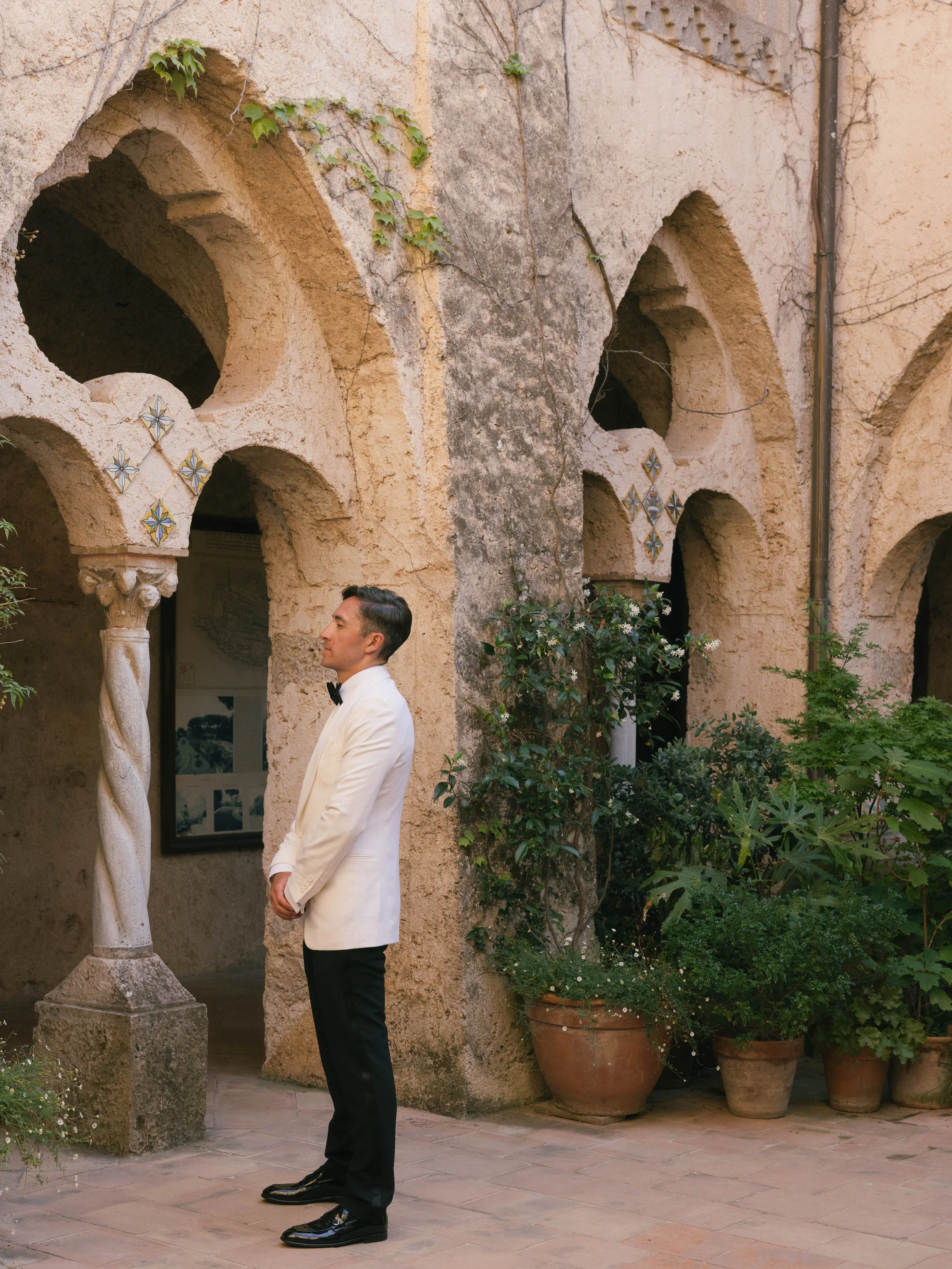 A man in a white tuxedo with a black bow tie and black shoes standing outdoors near a stone archway and potted plants against a beige wall with arches.