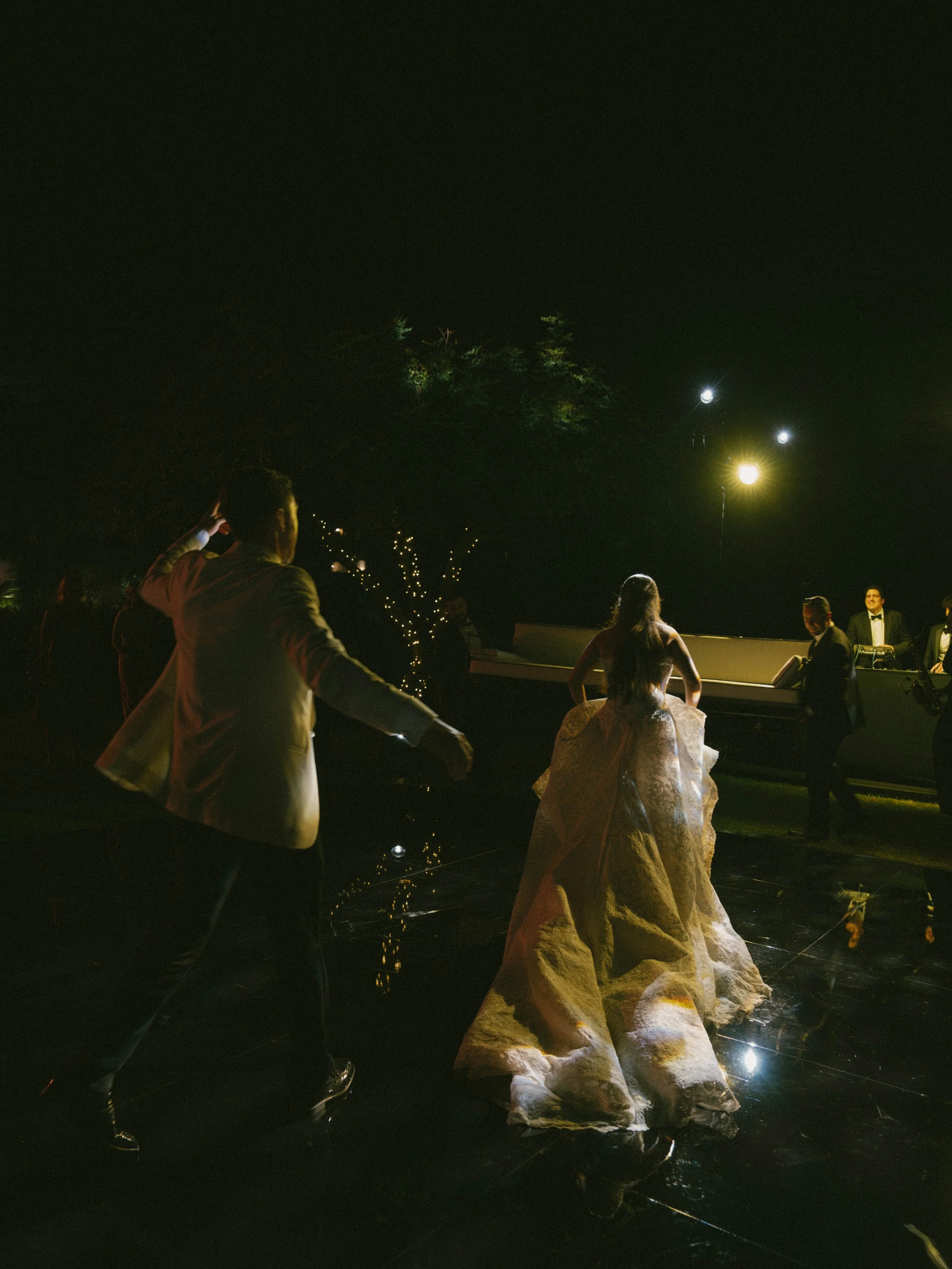 People dancing at a nighttime outdoor event with string lights, trees, and a DJ booth in the background.