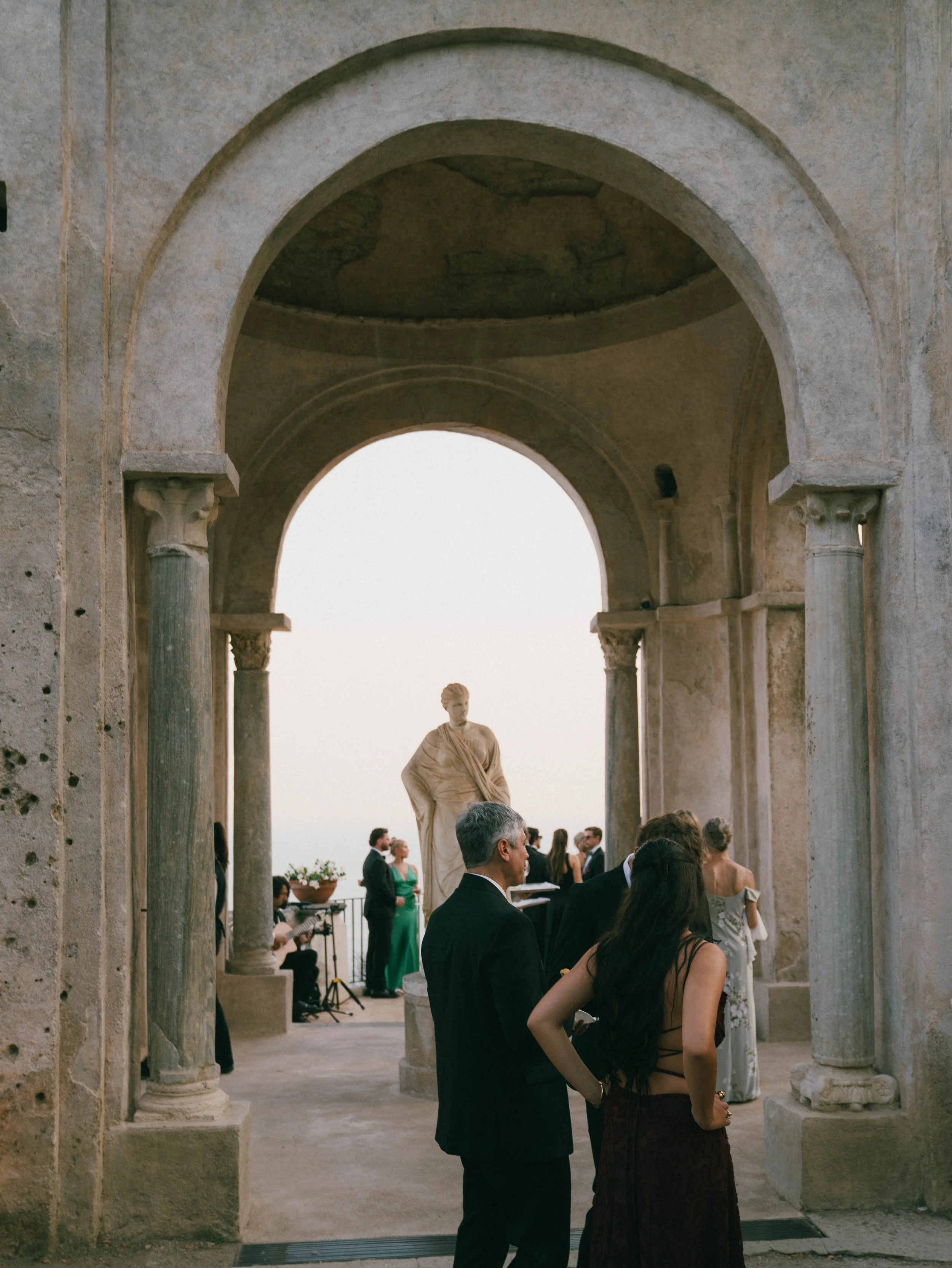 A wedding ceremony taking place on a terrace with classical architecture, including stone columns and arches, and a statue of a woman in the background. Several guests, dressed formally, are present, some standing and some seated, with musicians play