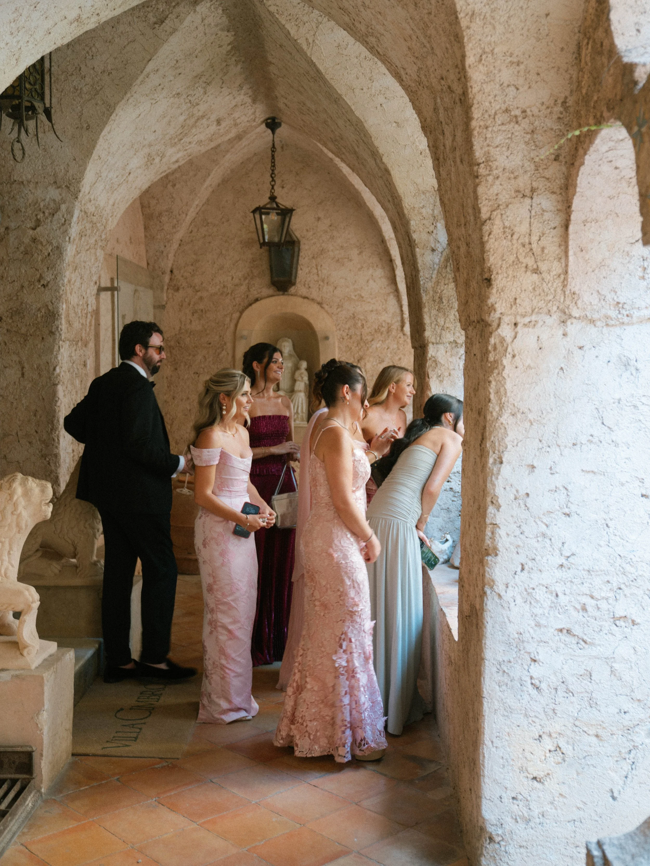 Group of people dressed in formal attire standing in an arched stone hallway, possibly a wedding or special event.