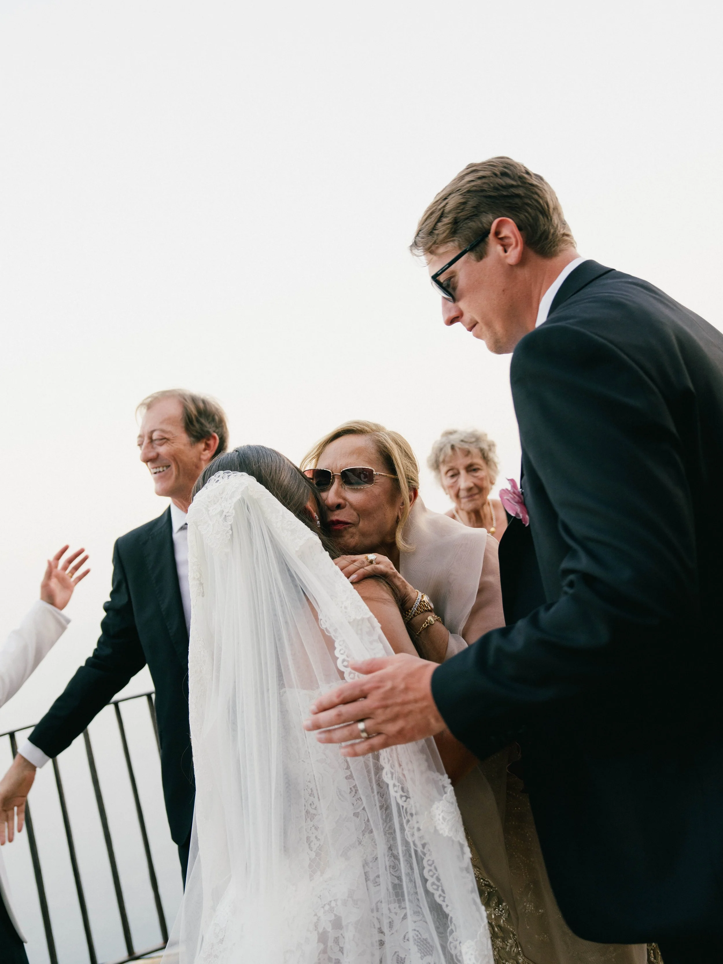 A group of people sharing a heartfelt moment at a wedding, with a woman in a white wedding dress embracing a man in glasses and a dark suit, surrounded by smiling older adults.