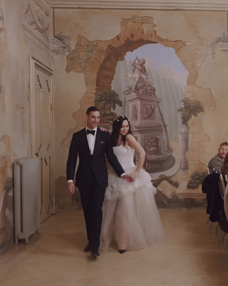 A bride and groom walking together at their wedding reception, with a painted mural of a fountain and a cherub behind them.