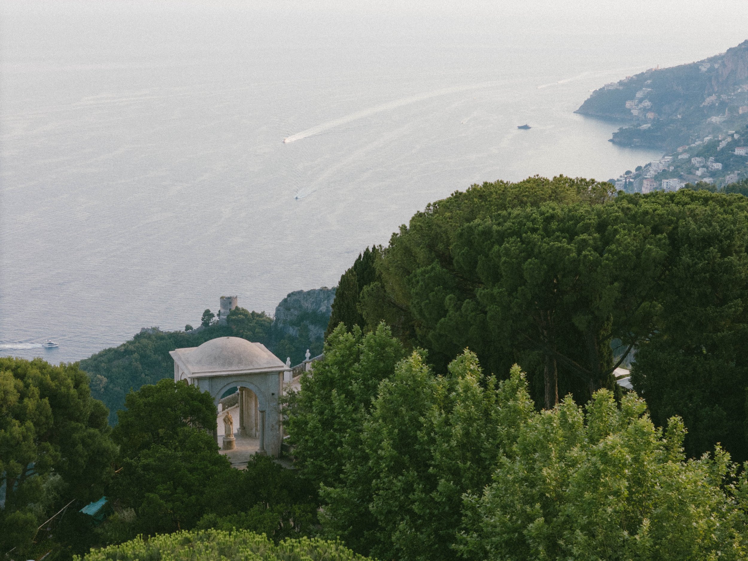 A scenic view of the Amalfi Coast with lush green trees, a white archway structure, and boats on the water in the distance.