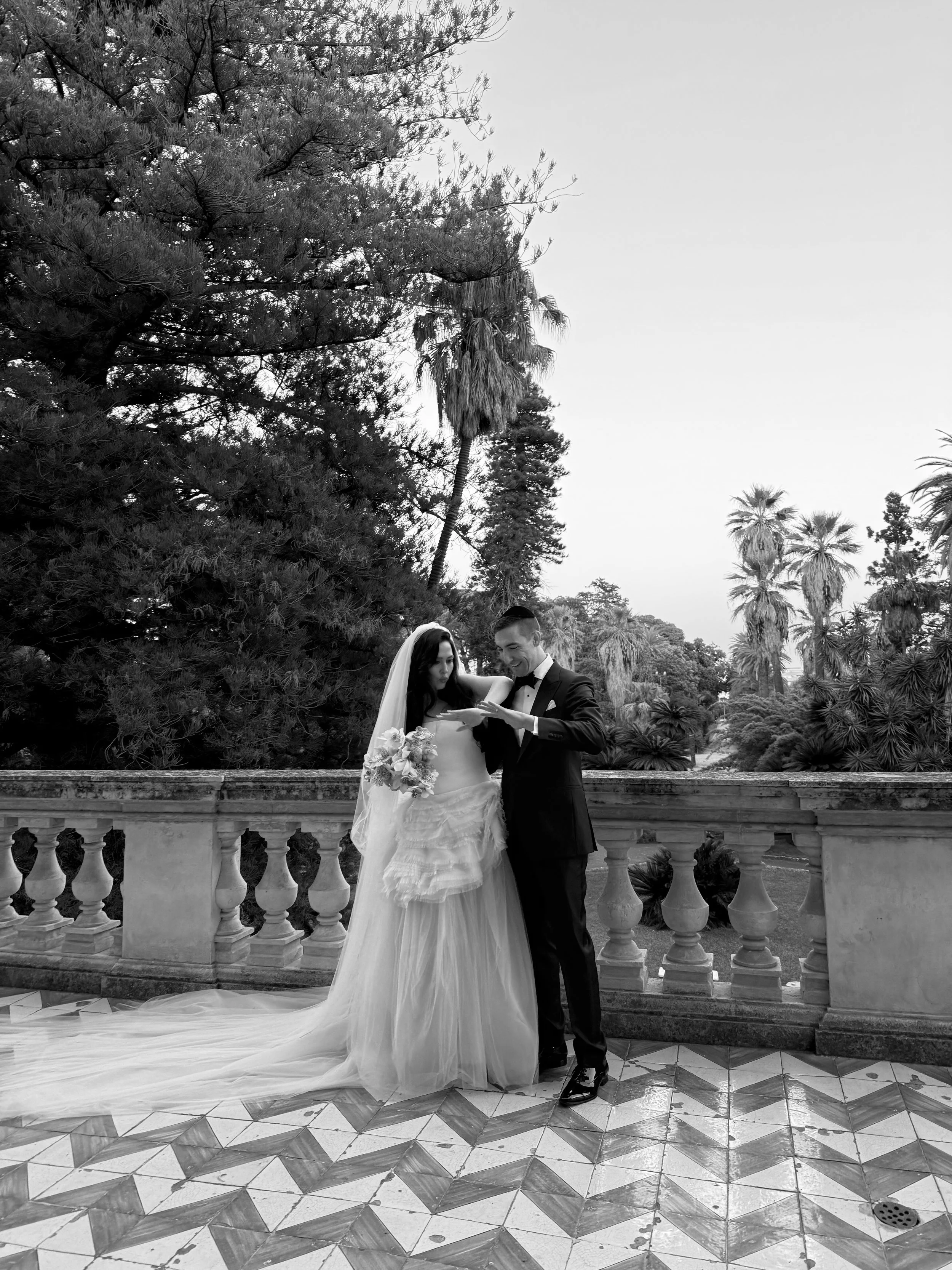 Black and white photo of a bride and groom standing on a balcony, with the bride holding a bouquet and dressed in a wedding gown, and the groom in a tuxedo, sharing a moment together outdoors with palm trees and lush greenery in the background.
