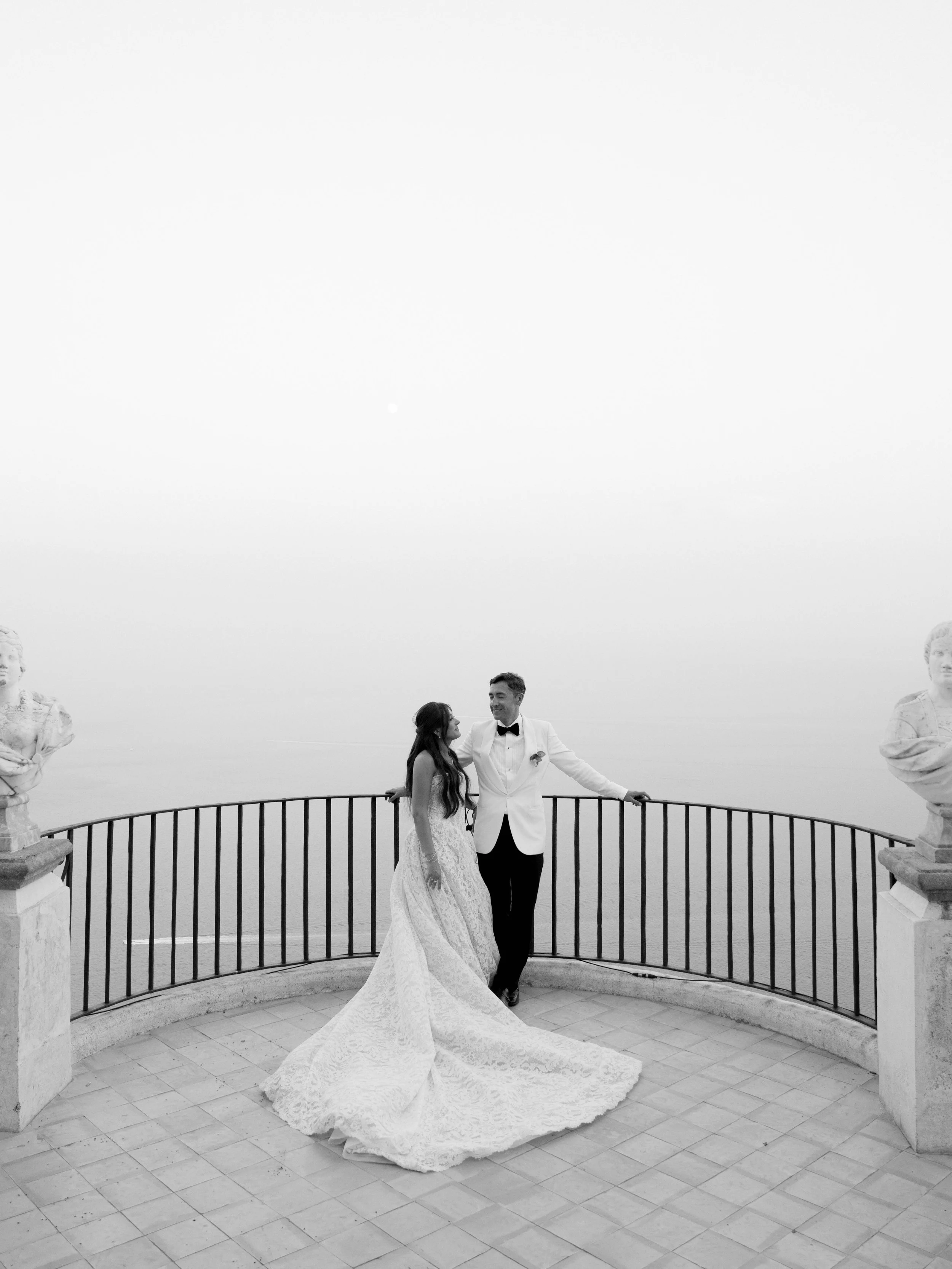 Black and white photo of a couple in wedding attire standing on a balcony, embracing, with a woman in a wedding gown with a long train and a man in a tuxedo, surrounded by statues and a balustrade, overlooking a misty or cloudy sky.