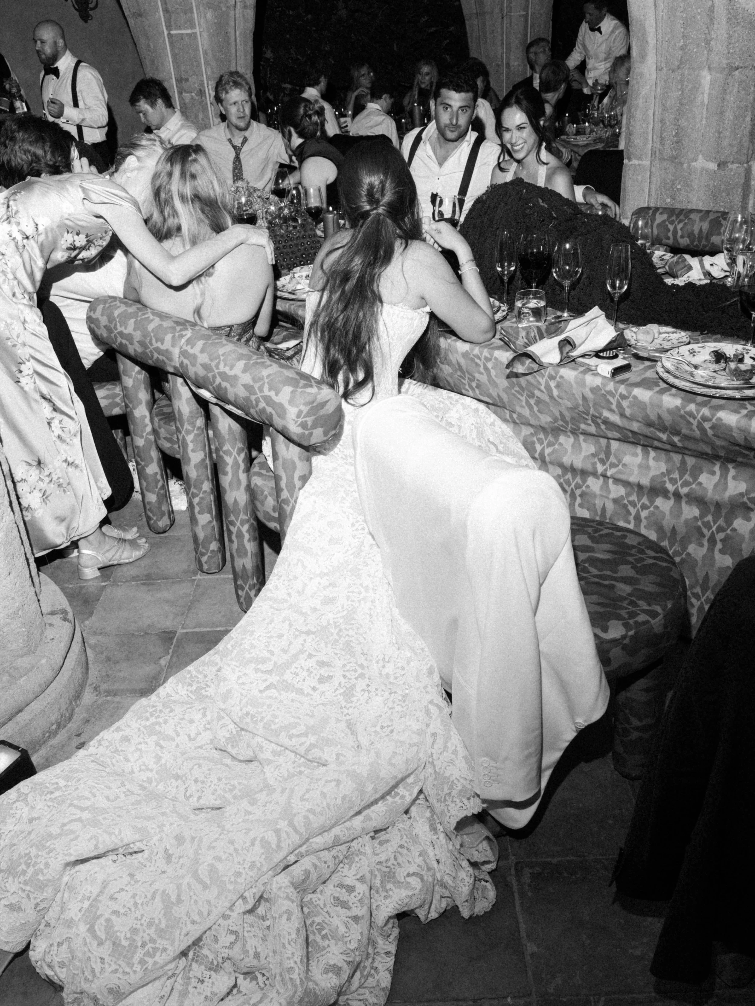 A black-and-white photo of a lively social gathering at a restaurant or banquet hall, with people sitting at tables, exchanging conversations, and enjoying food and drinks. The scene shows a variety of people, some smiling and engaged with each other