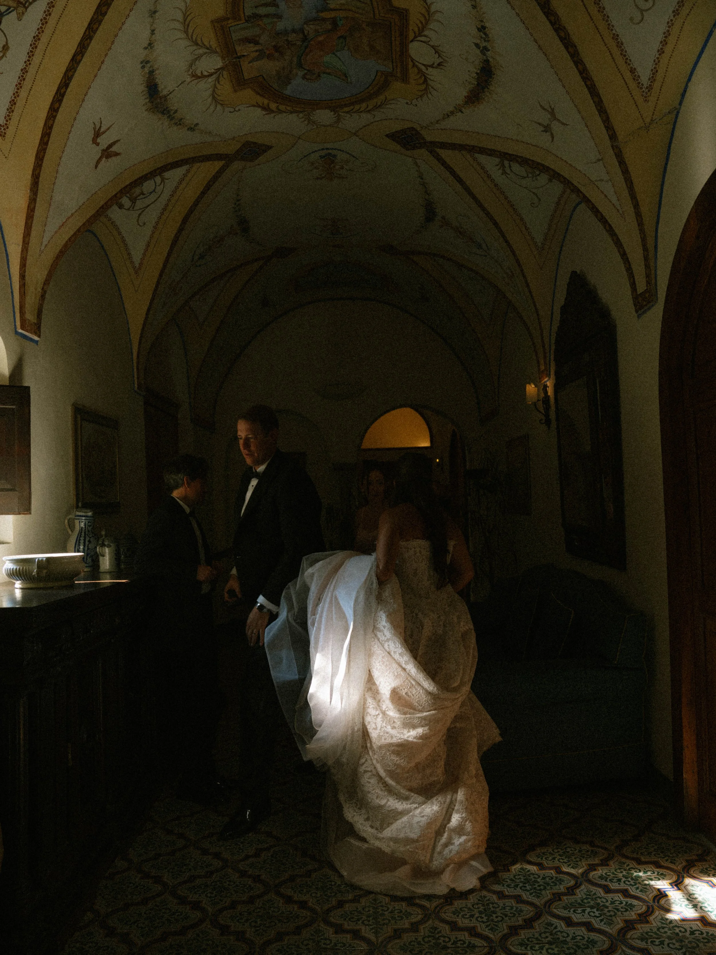A bride in a white wedding dress with a veil is getting ready, assisted by a woman in a silver dress, in a dimly lit hallway with ornate ceiling art and patterned tile floors. Two men in tuxedos stand nearby.