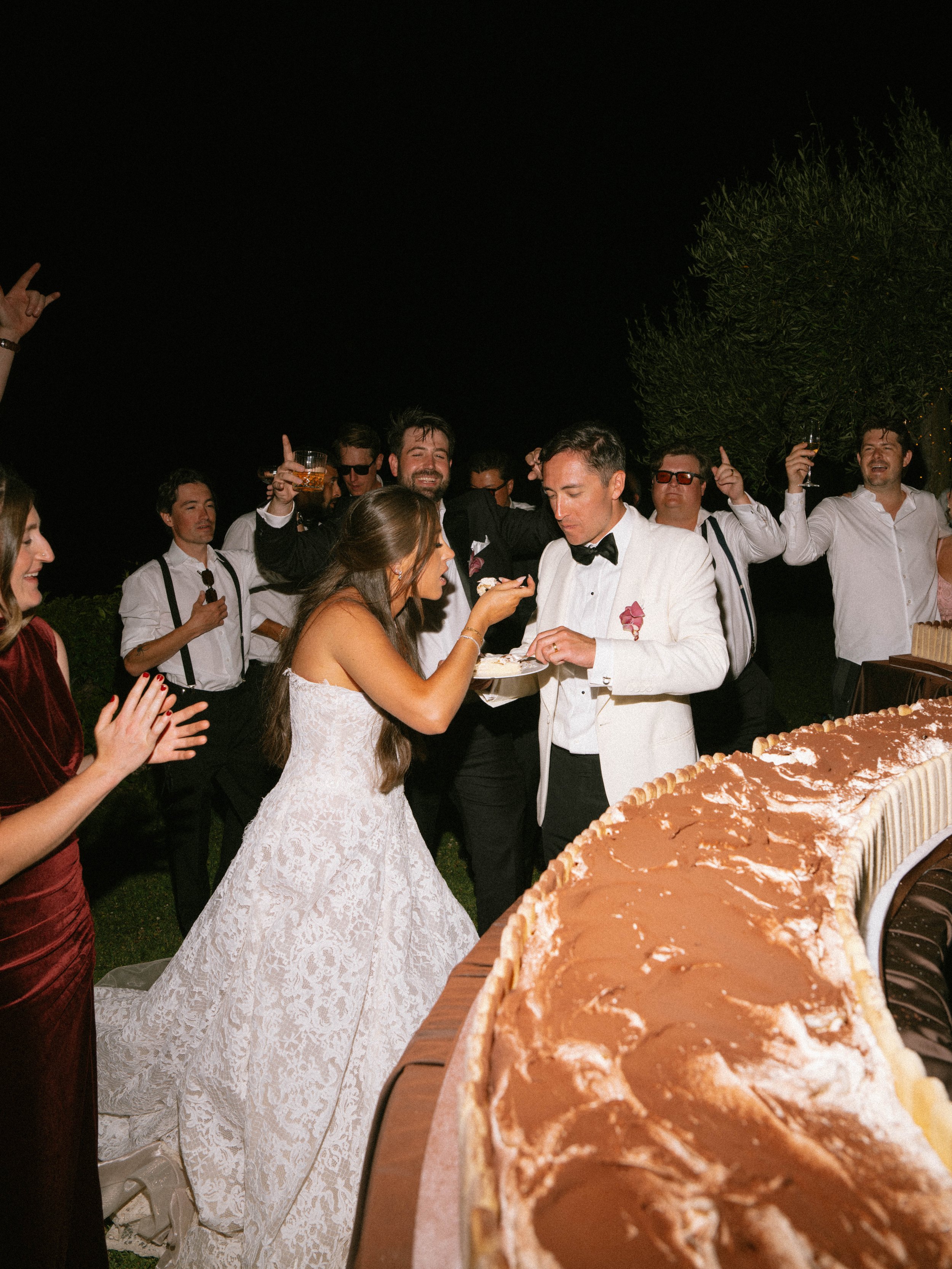 A group of people, including a bride in a white lace wedding dress and a groom in a white tuxedo, are celebrating outdoors at night with a large cake, some holding drinks, and smiling happily.