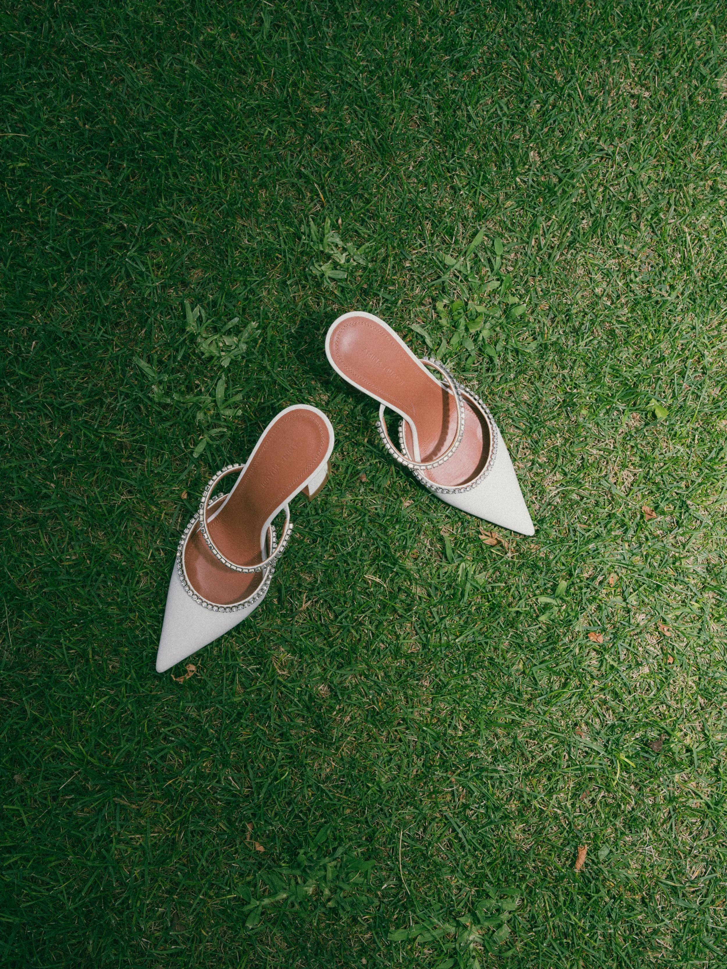 A pair of white high-heeled shoes with pointed toes decorated with rhinestones, placed on green grass.